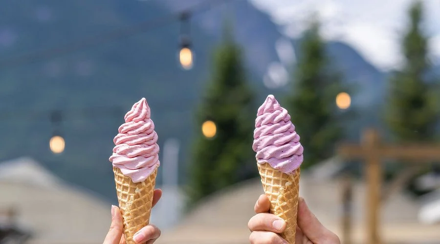 Two hands holding ice cream cones with pink and purple ice cream against an outdoor background with trees and string lights.