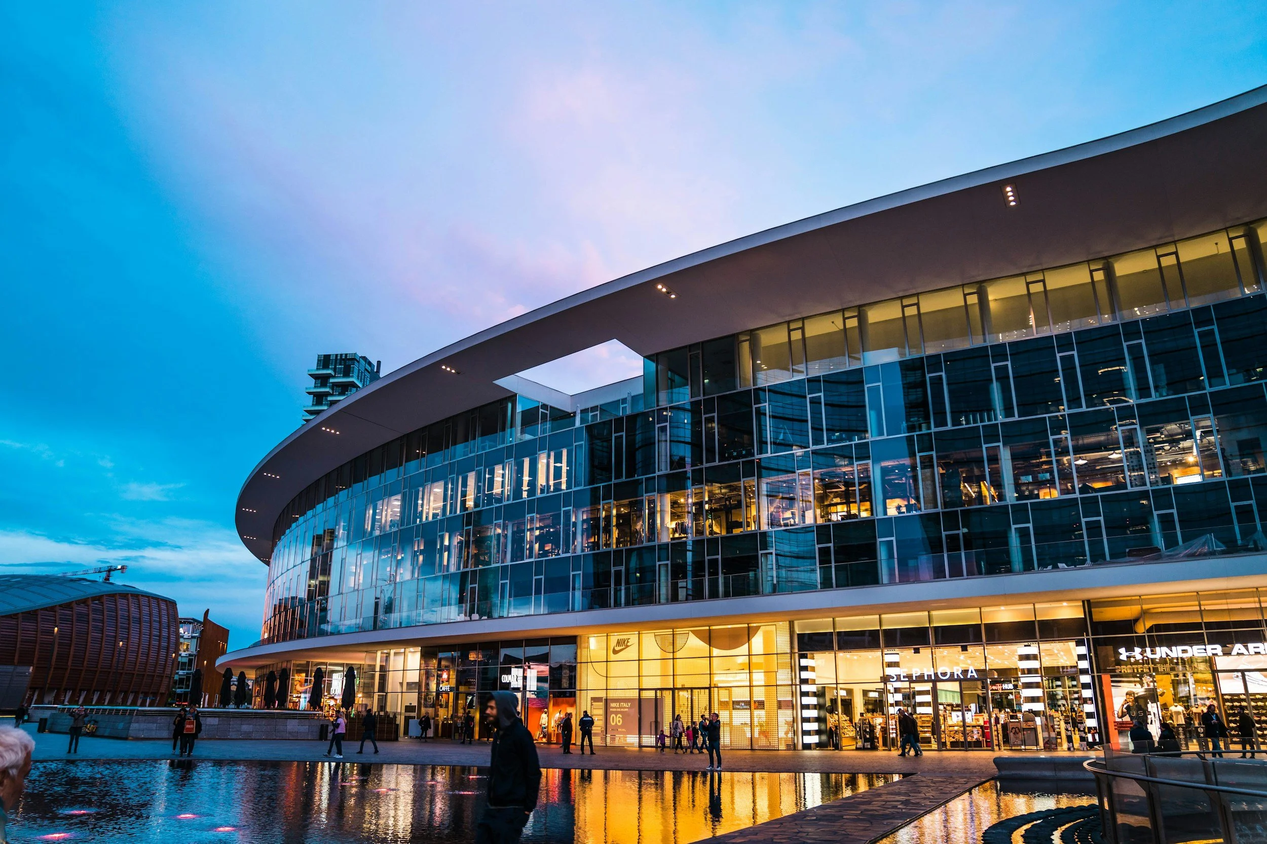 Modern glass-fronted shopping mall with illuminated store signs, including Nike, Sephora, and Under Armour, during evening with people walking outside and reflections in water.