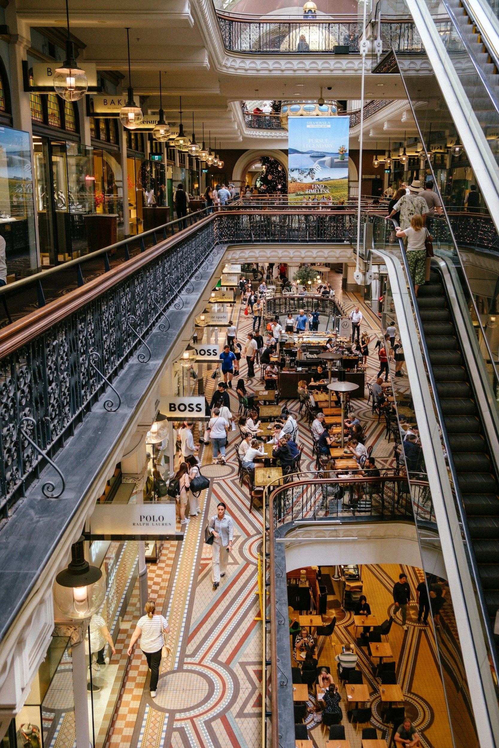 Multiple floors of a shopping mall with people walking, shopping, and sitting at cafes; ornate decor, escalators, and storefronts visible.