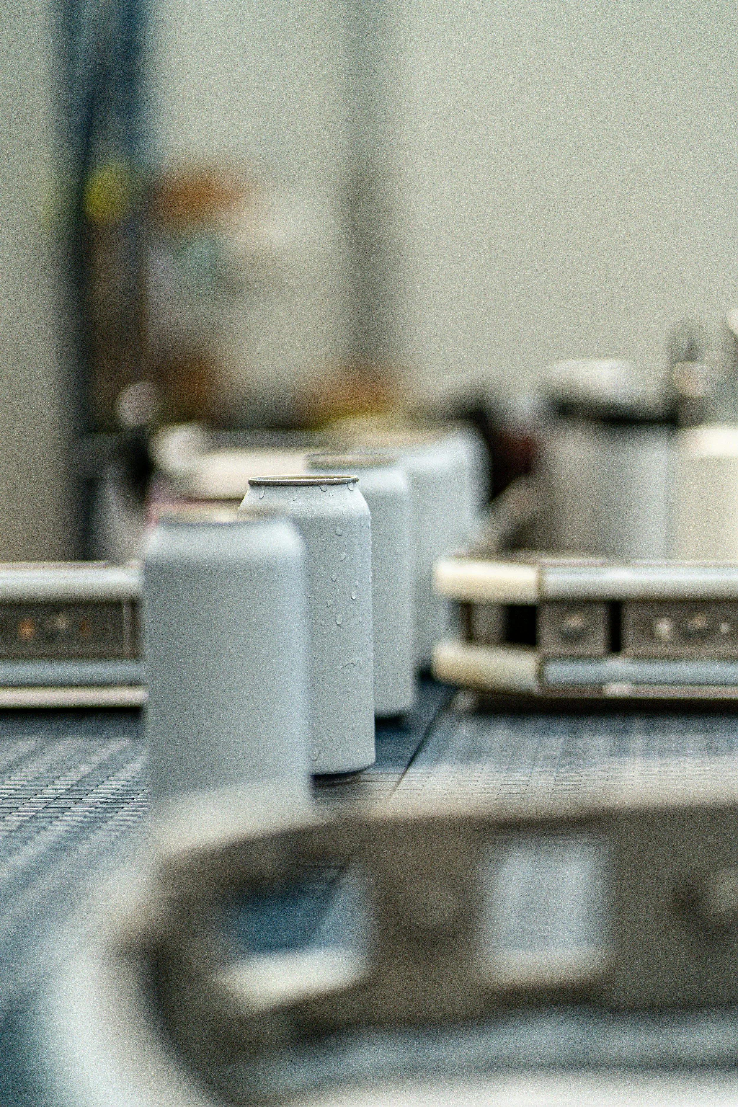 Close-up of three aluminum cans with water droplets on them, placed on a metallic countertop in a kitchen or break room, with blurred appliances and objects in the background.