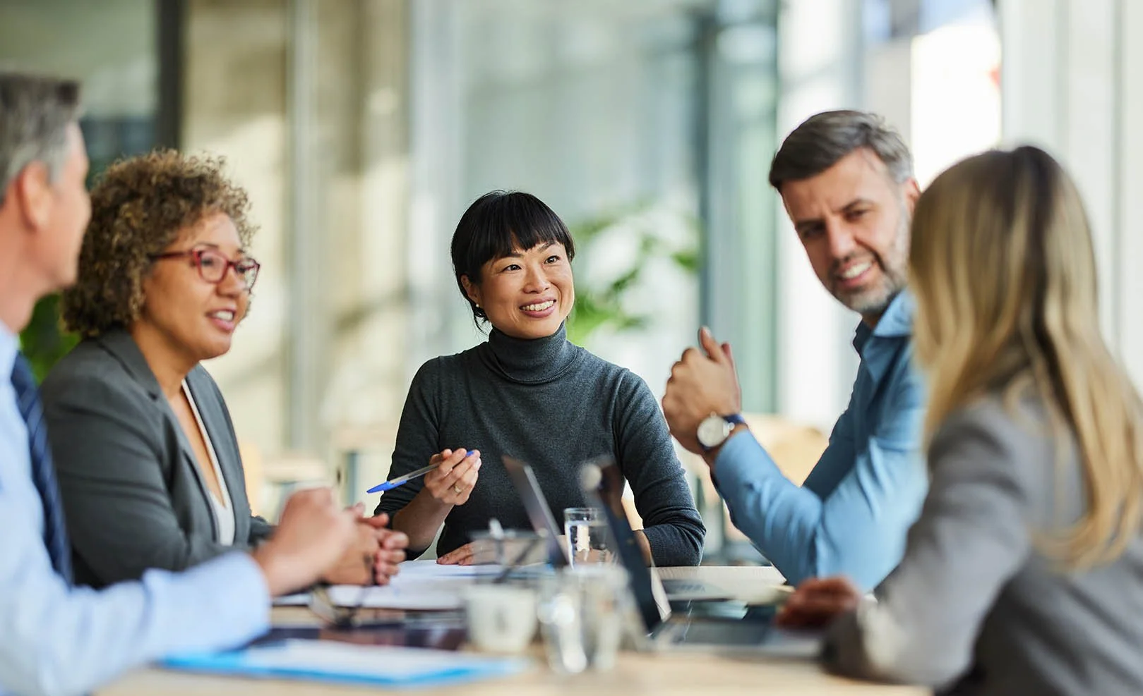 Group of five diverse professionals having a discussion at a business meeting in a modern office with large windows and natural light.