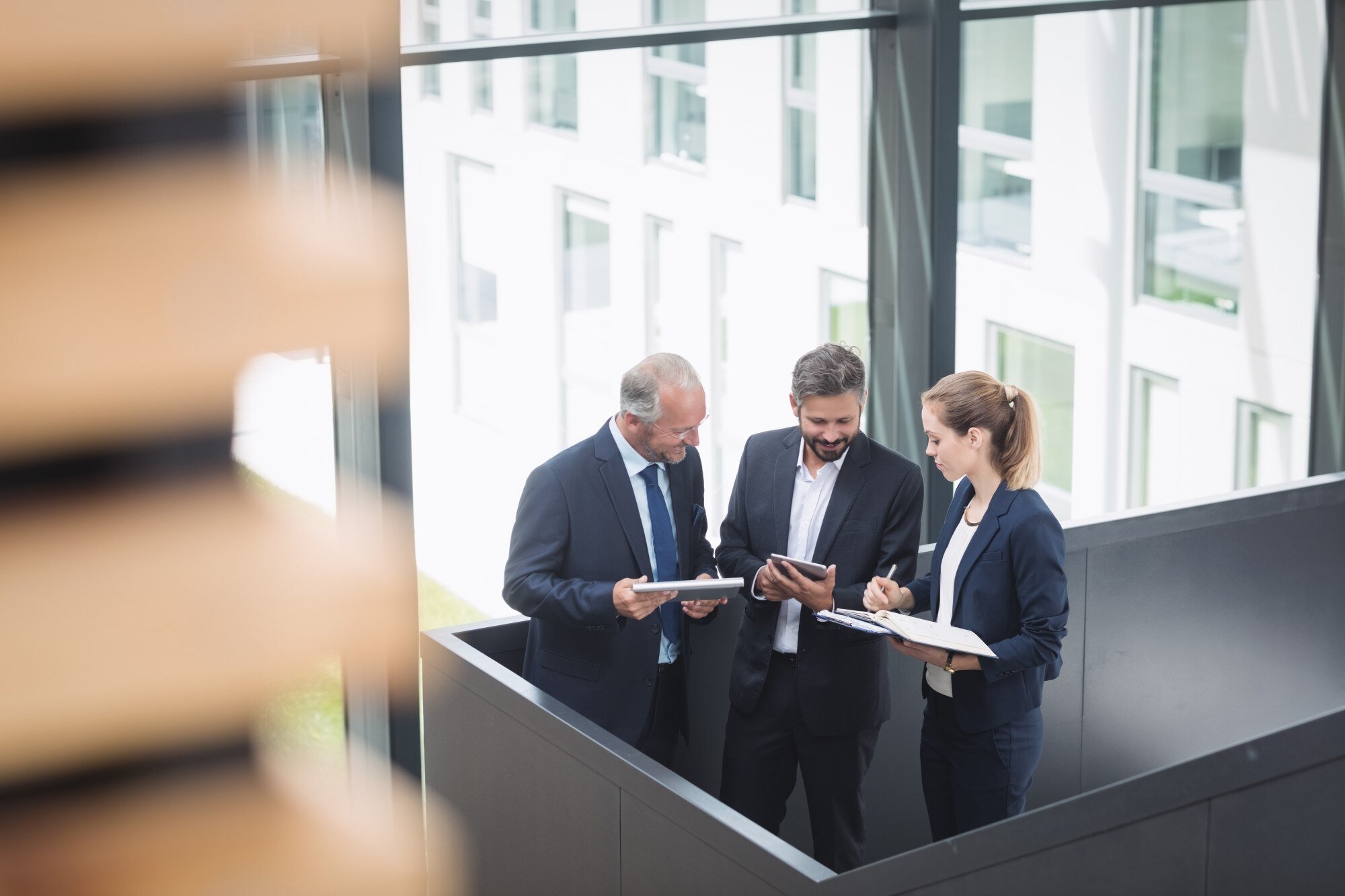 Three business professionals, two men and one woman, are having a discussion, looking at a tablet, in a modern office building with large windows.