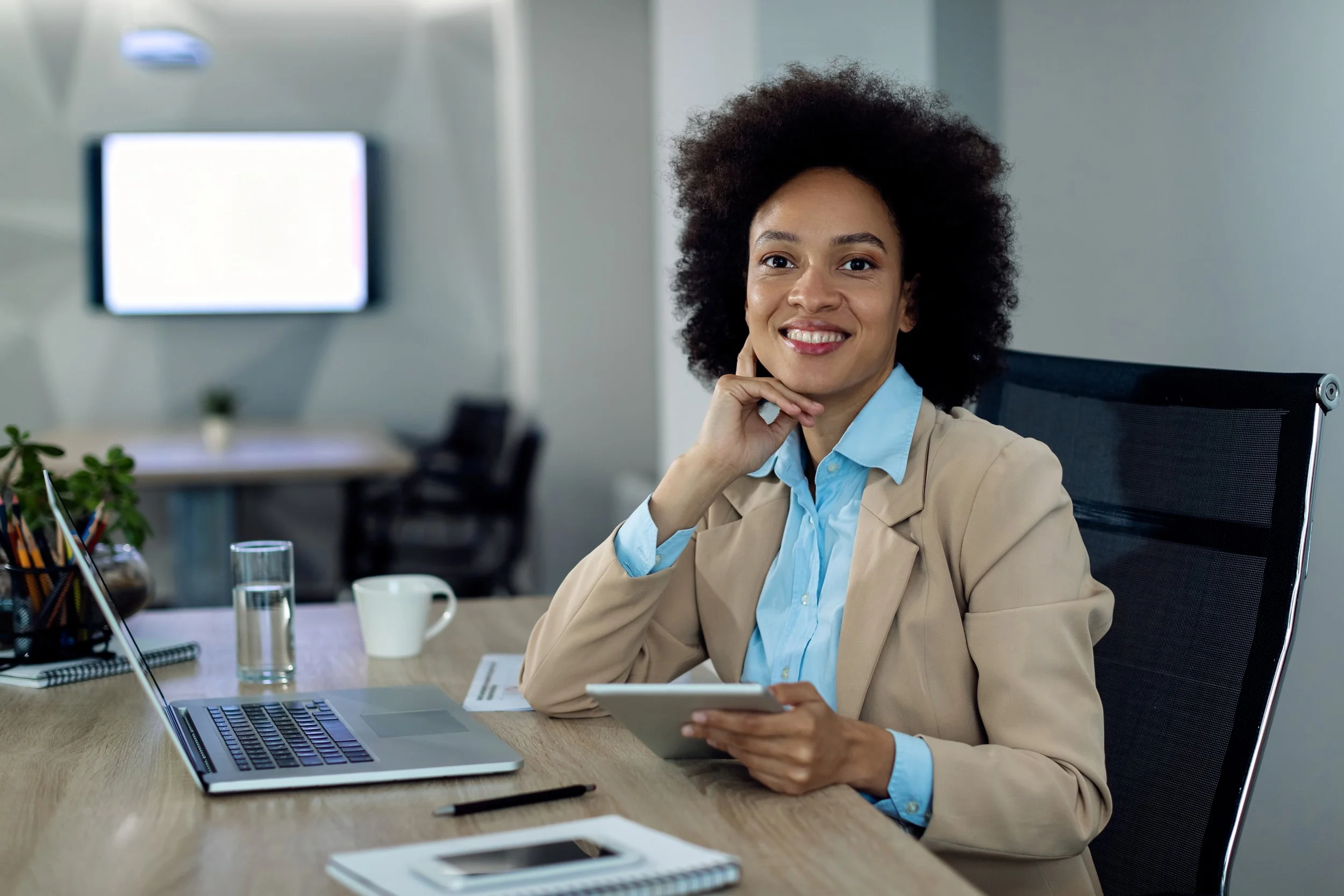 Professional woman sitting at a desk with a laptop, tablet, and notebook, smiling in an office setting.