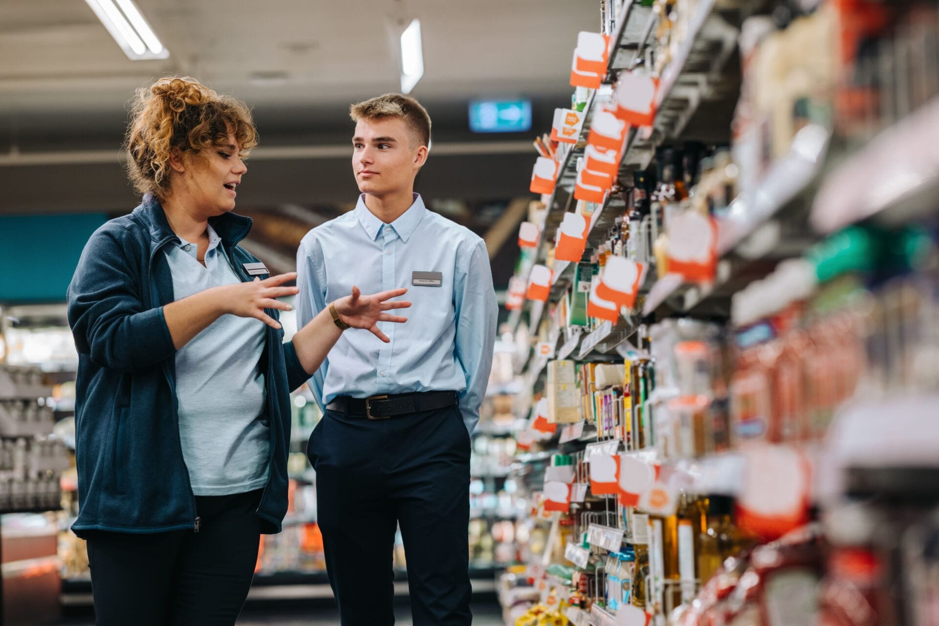 Two store employees, a woman and a man, are having a conversation in an aisle of a grocery store with shelves stocked with various food products.