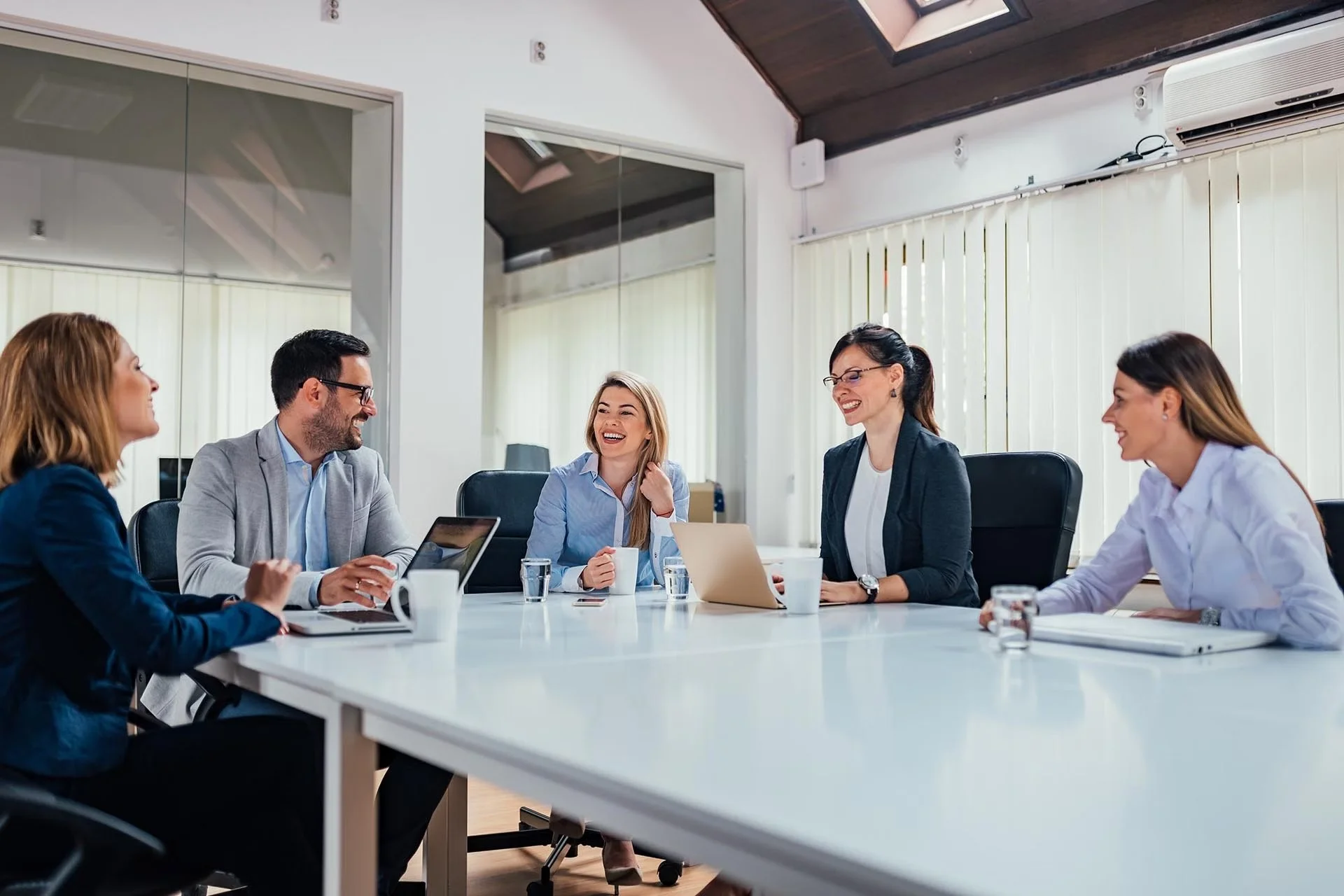 Five professionals sitting around a white conference table in a modern office, engaged in a discussion, smiling, with laptops, tablets, notepads, and cups on the table.