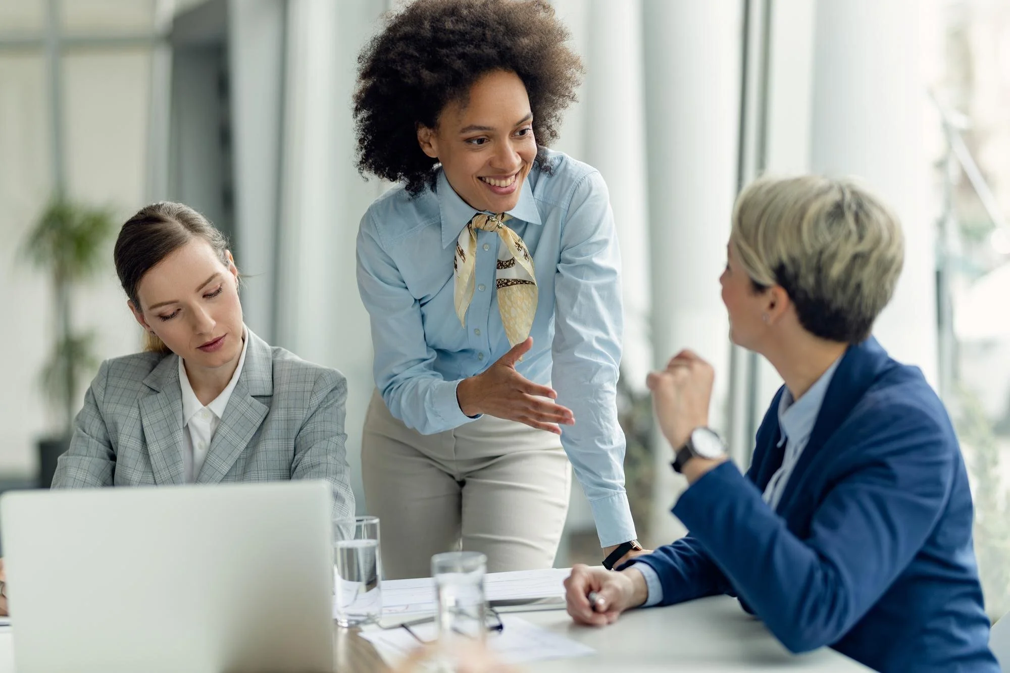 Three women in a business meeting. One woman is standing, smiling, and talking to a woman with short blonde hair who is sitting at a table, while the third woman is seated, looking down. The table has glasses of water, papers, and a laptop.