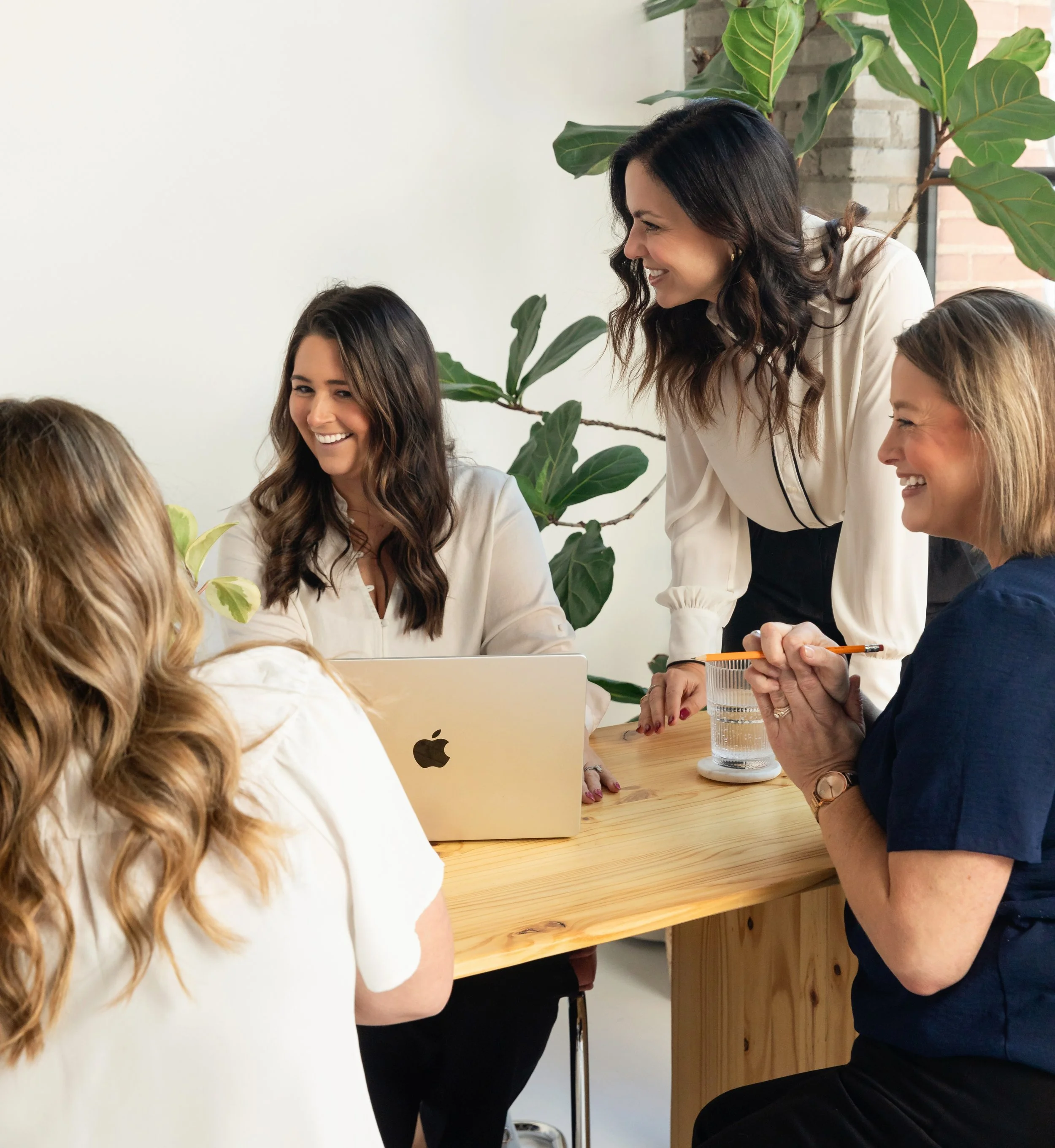 Four women in a meeting or discussion in a bright office with green plants, sitting around a wooden table with a laptop and a glass of water.