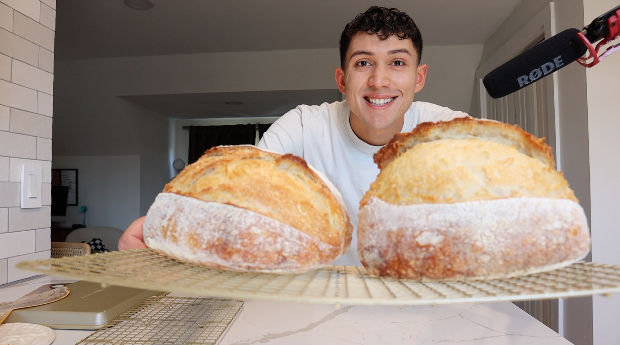 Young man smiling in a modern kitchen, showcasing two large loaves of sourdough bread on a cooling rack.