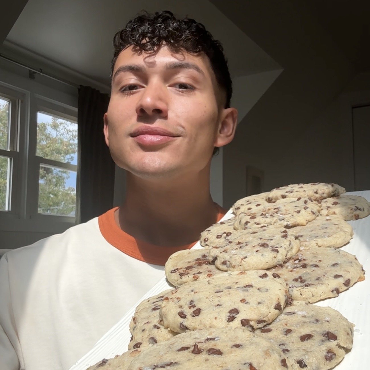 A young man with curly dark hair holding a tray of chocolate chip cookies, standing indoors near a window.