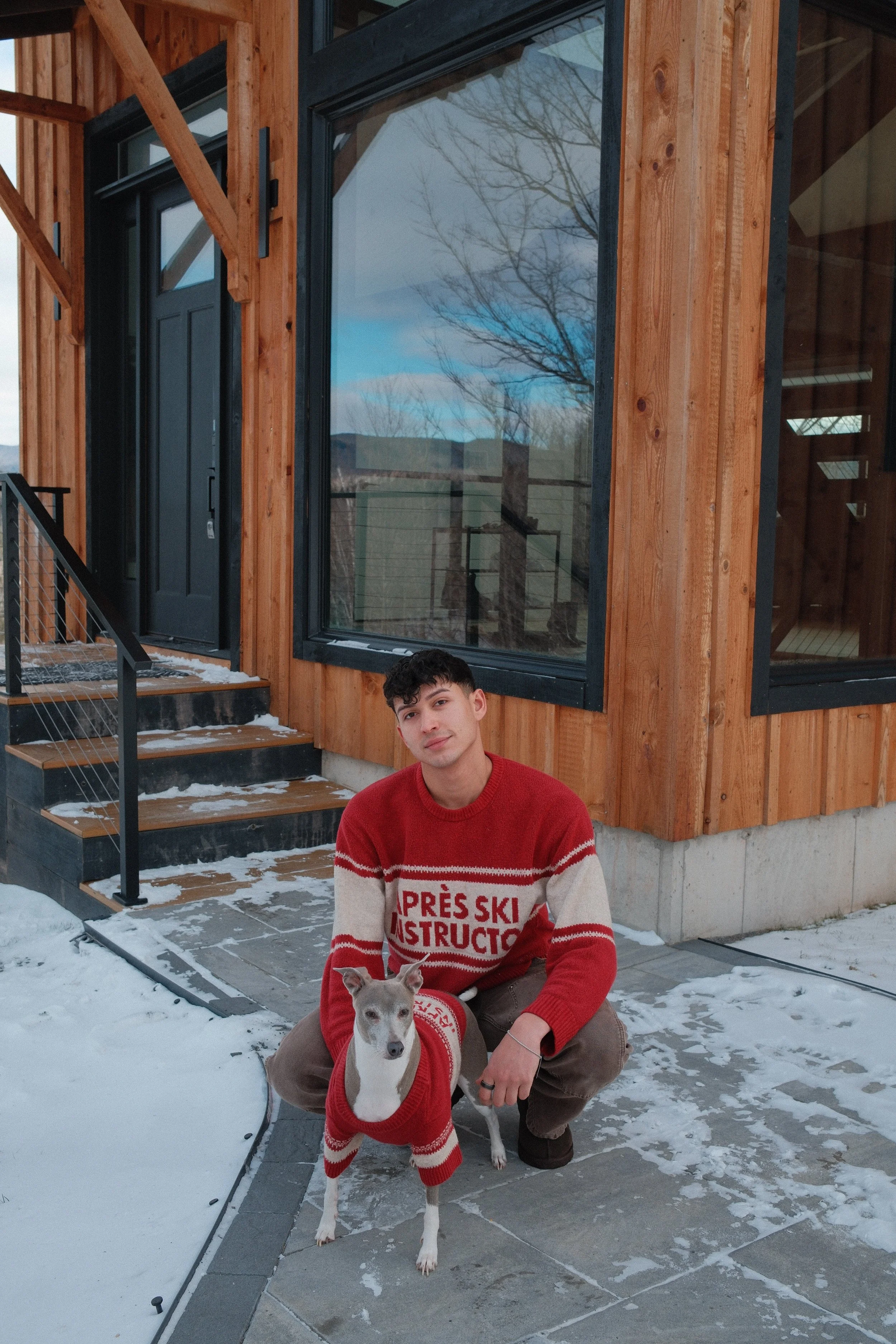 A young man in a red sweater with white and red text that reads 'Après Ski Instructor' crouches beside a dog in matching sweater outside a wooden house with large windows, snow on the ground, and leafless trees reflected in the windows.