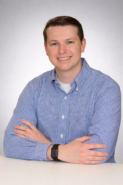 Portrait of a young man with short brown hair, wearing a blue checkered shirt, smiling, with arms crossed, against a plain gray background.