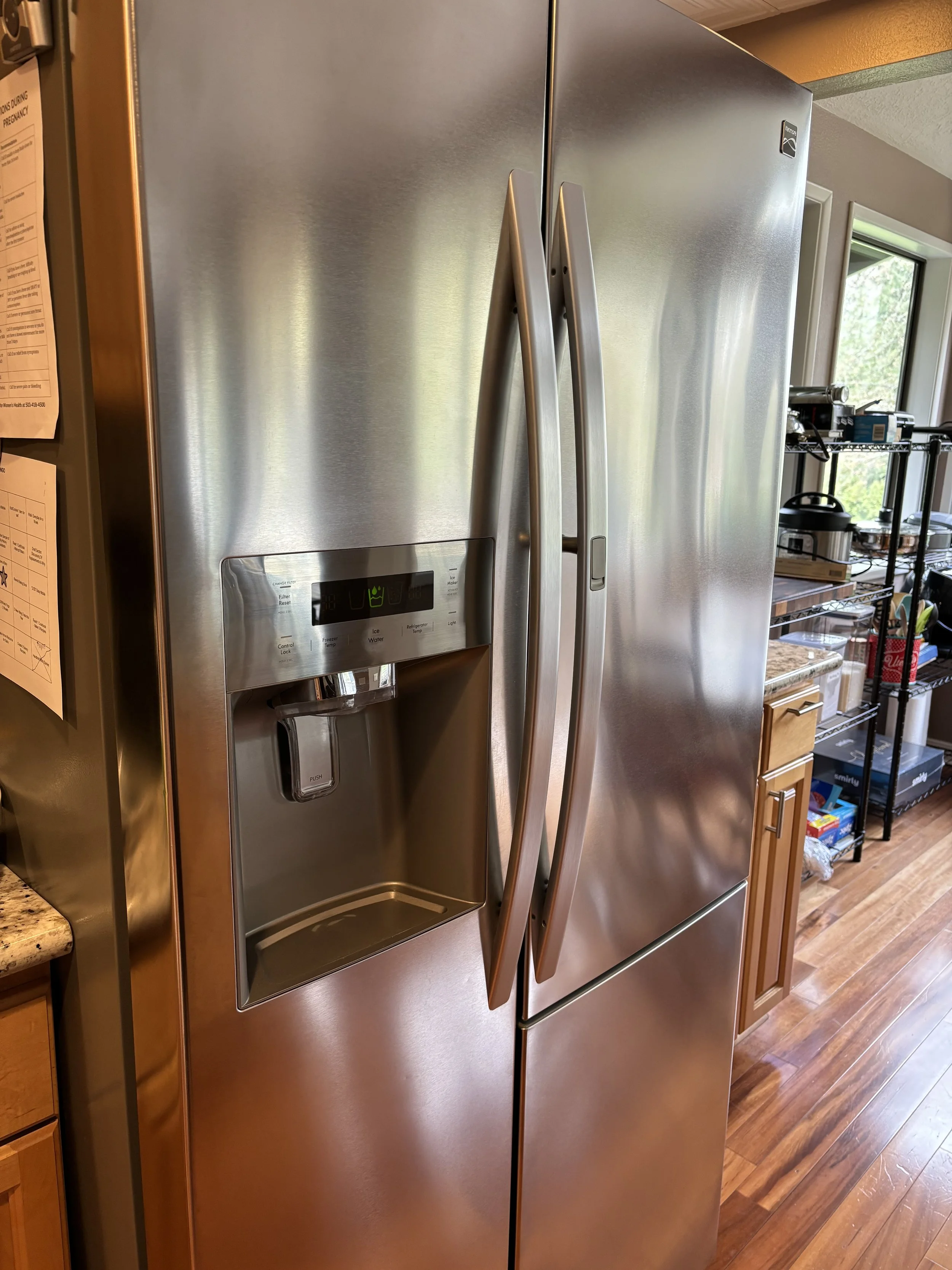 Stainless steel side-by-side refrigerator with a water and ice dispenser in a kitchen with wooden cabinets and a wire shelving unit.