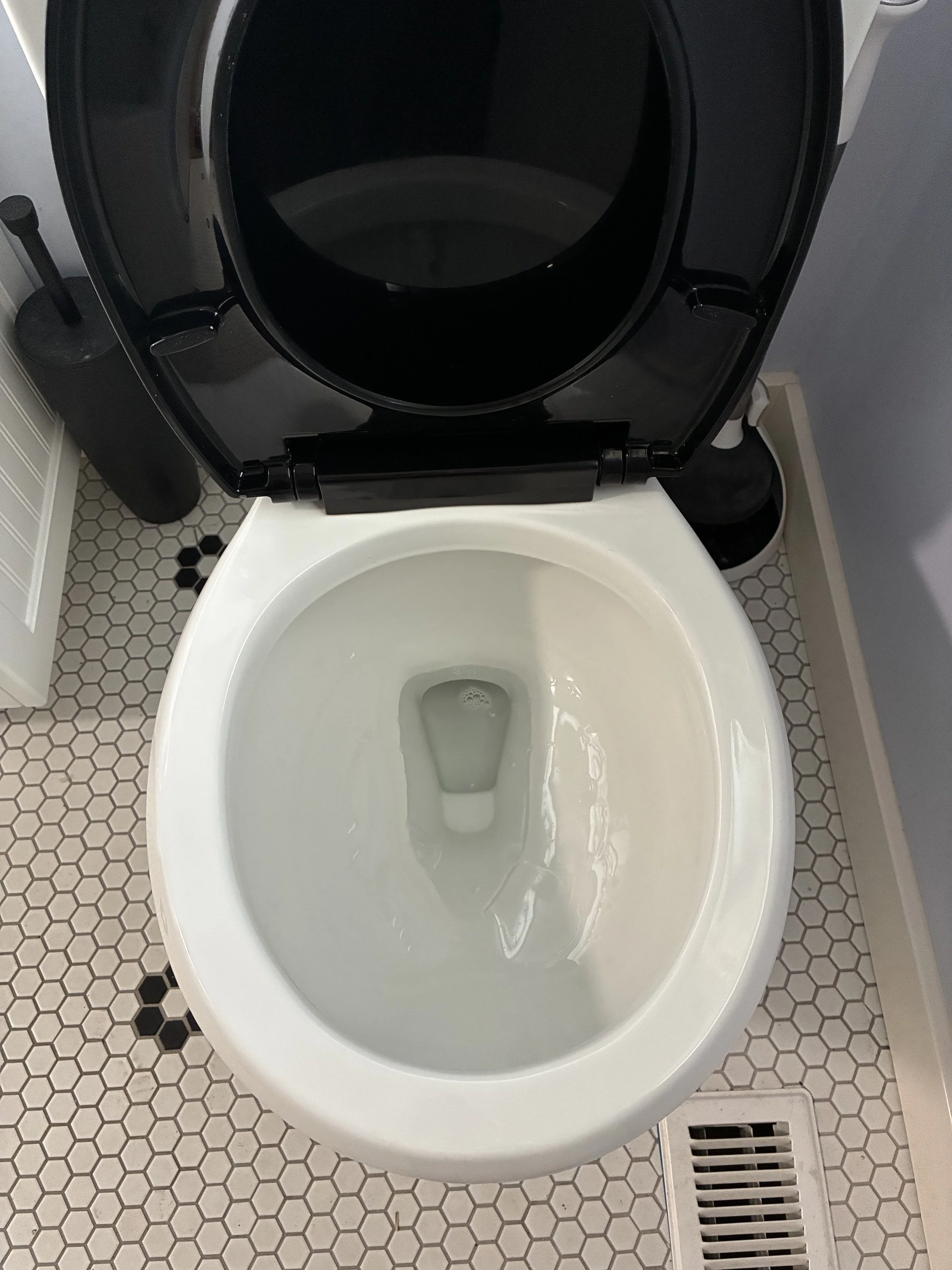 Open toilet with water inside, in a bathroom with hexagon tile flooring and a vent near the bottom of the image.