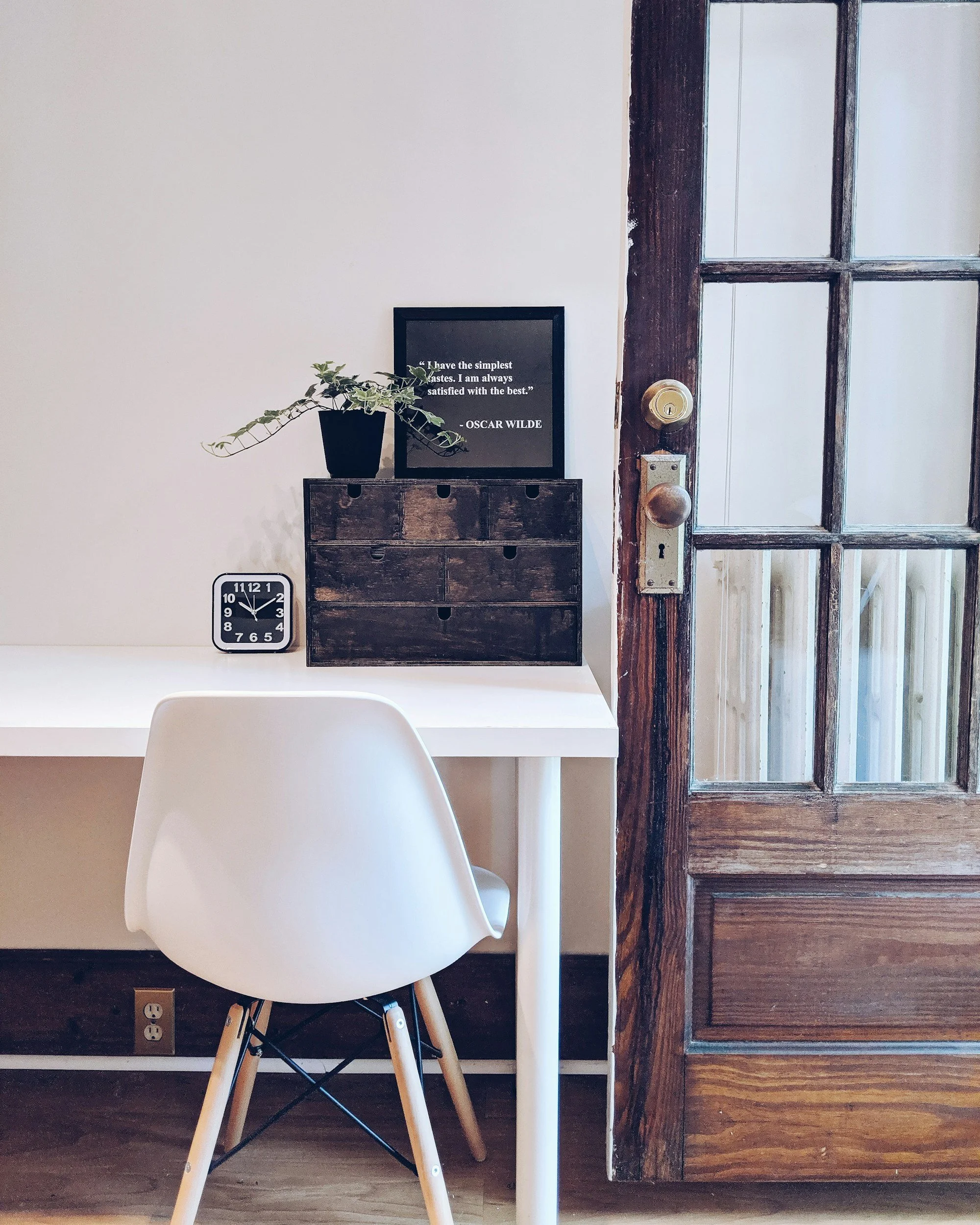 A white desk with a white chair, a black clock, a small black storage box, a potted plant, and a framed quote on a wall, next to a wooden door with glass panels.