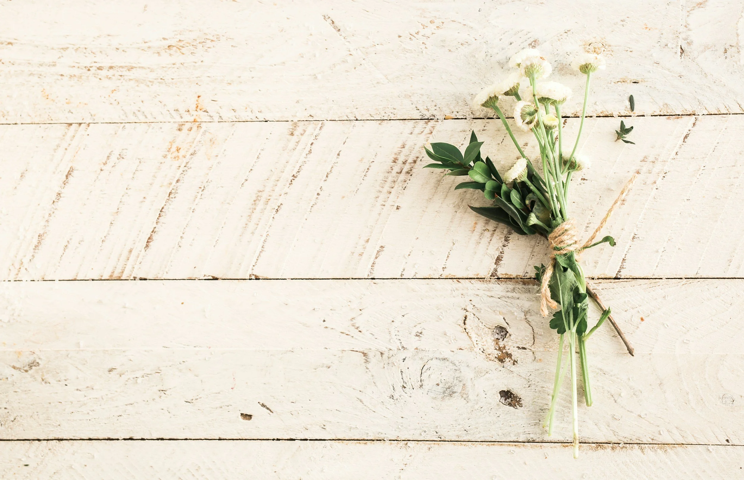 A small bouquet of white flowers and green leaves tied with twine, placed on a rustic white wooden surface.