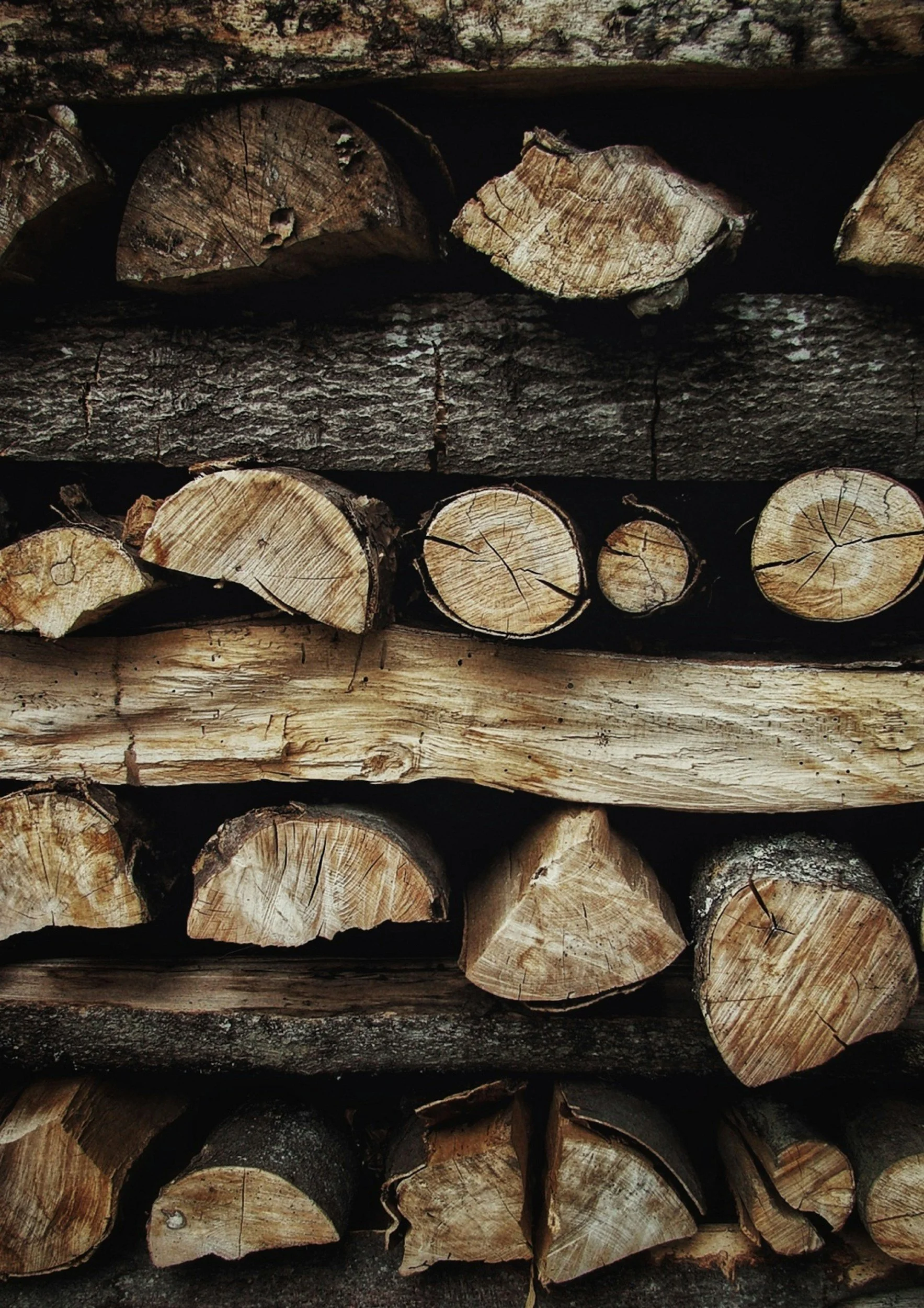 Stacked firewood logs with visible cut ends, some with cracks and bark, arranged horizontally in a pile.