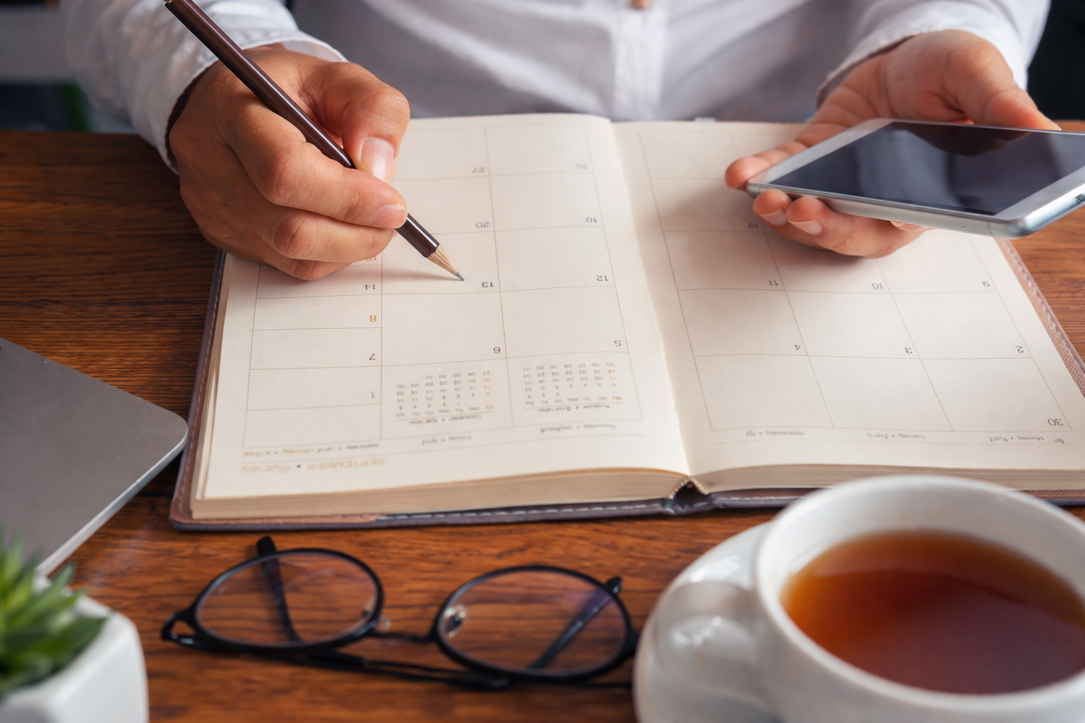 Person using a pen and smartphone at a desk with a planner, coffee, glasses, and a plant.