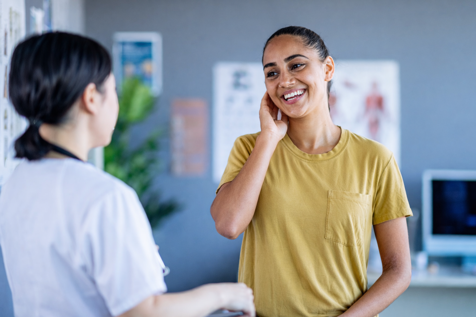 A young woman smiling and touching her face while talking to a healthcare professional in a medical office.