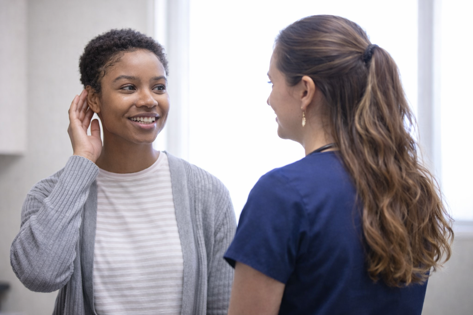 A nurse or healthcare worker smiling and talking to a woman in a clinical setting, with both women facing each other.