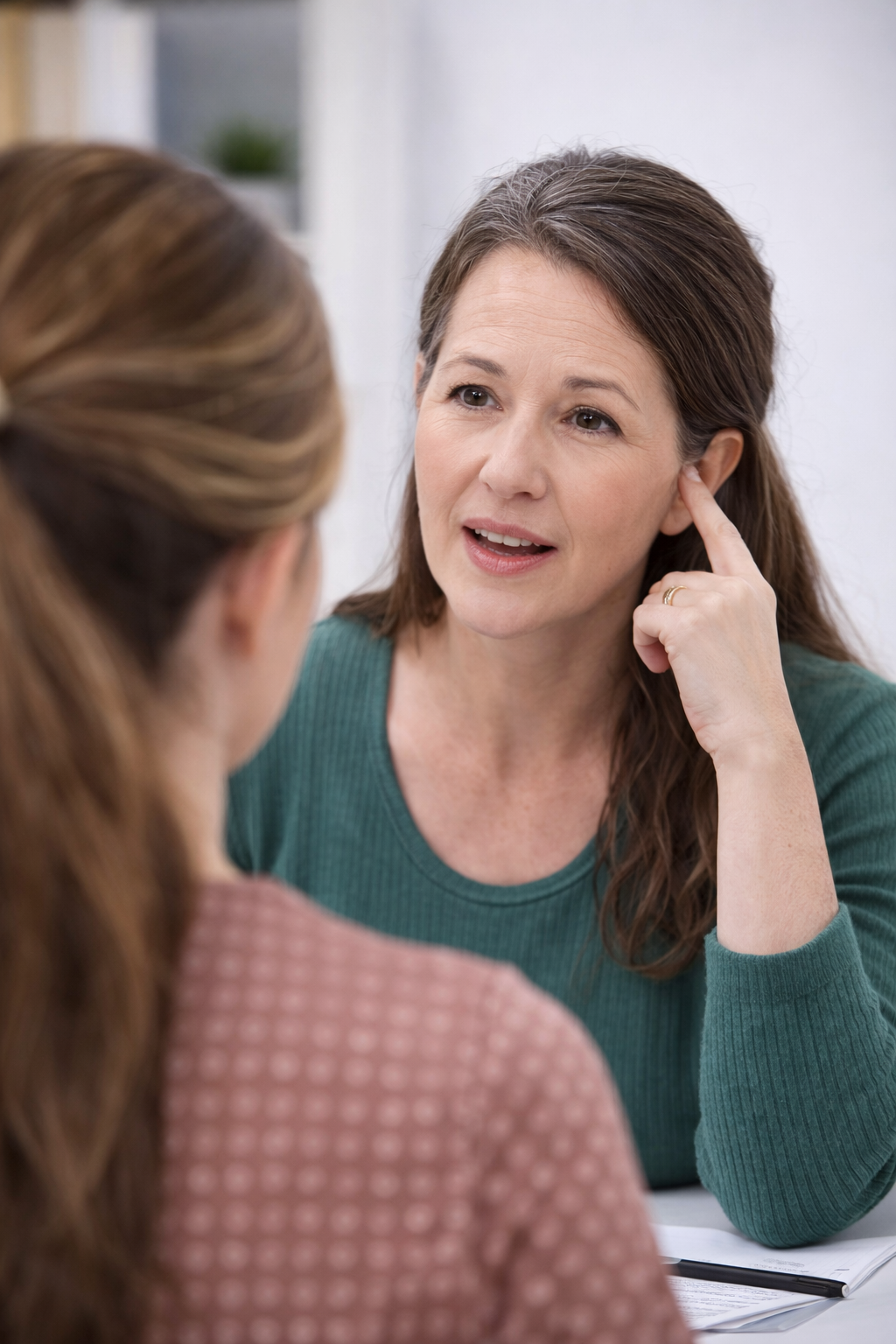 A woman with brown hair listening to another woman with brown hair, wearing a green sweater, in a conversation at a table.
