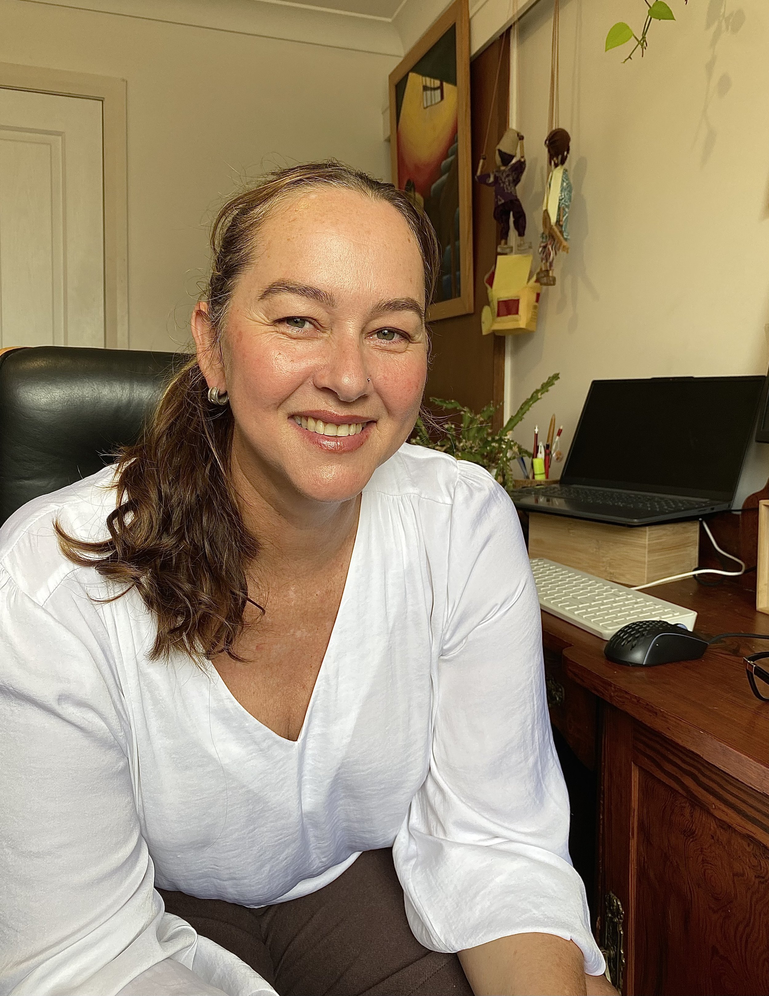 A woman with long, wavy brown hair and light skin smiling while sitting at a desk in a home office. She is wearing a white blouse and earrings.