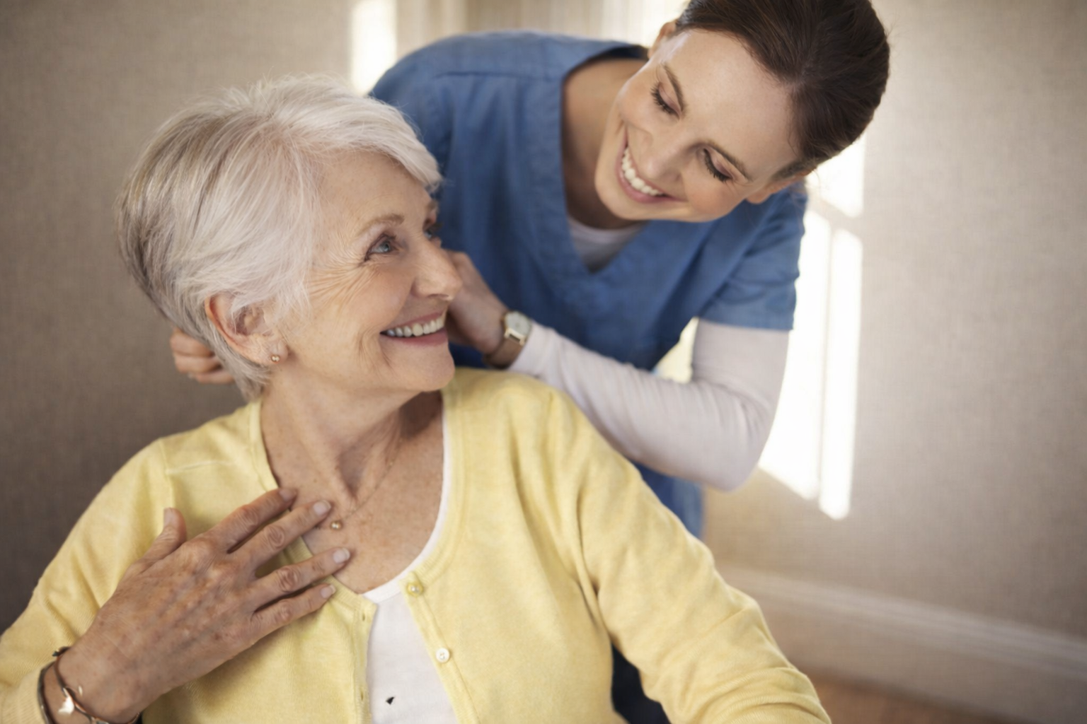 A young female healthcare worker in blue scrubs smiling and talking to an elderly woman with white hair, wearing a yellow cardigan, who is sitting and smiling back.