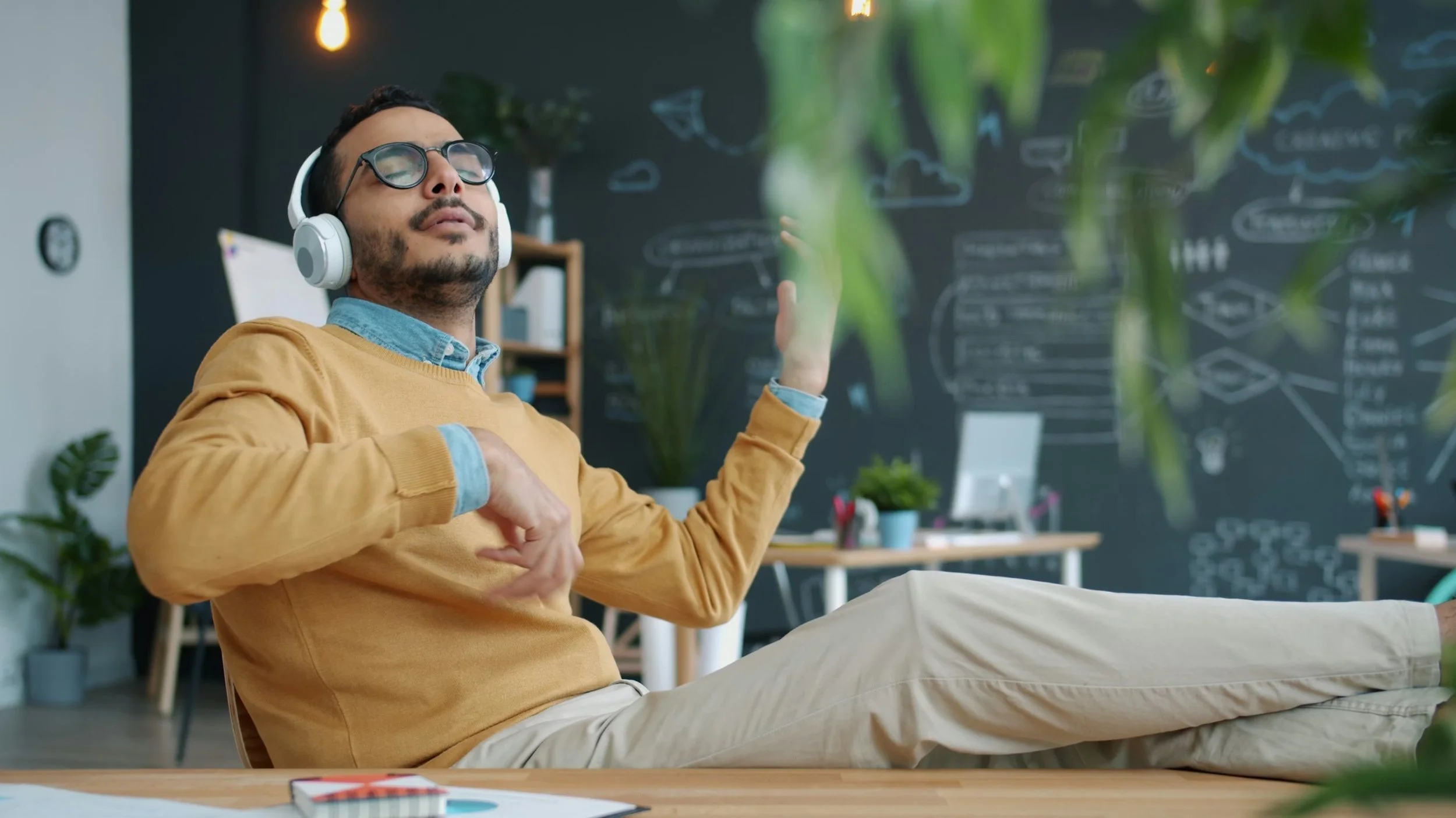 A man in a tan sweater and glasses is sitting in an office with his eyes closed and wearing headphones, appearing relaxed and enjoying music or meditation.