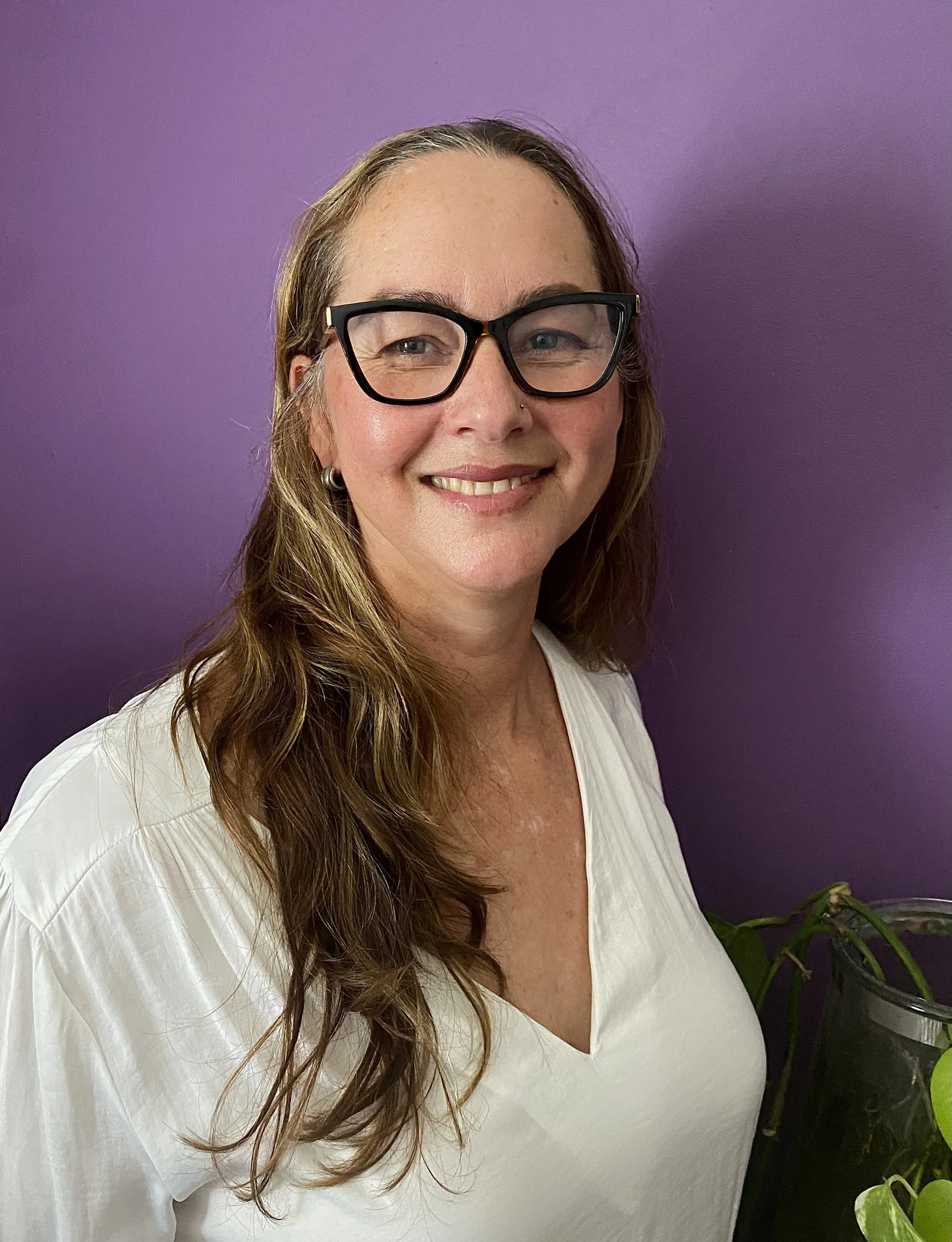 A woman with long wavy hair wearing glasses and a white blouse stands against a purple wall, smiling at the camera, with a potted plant visible on the right side.