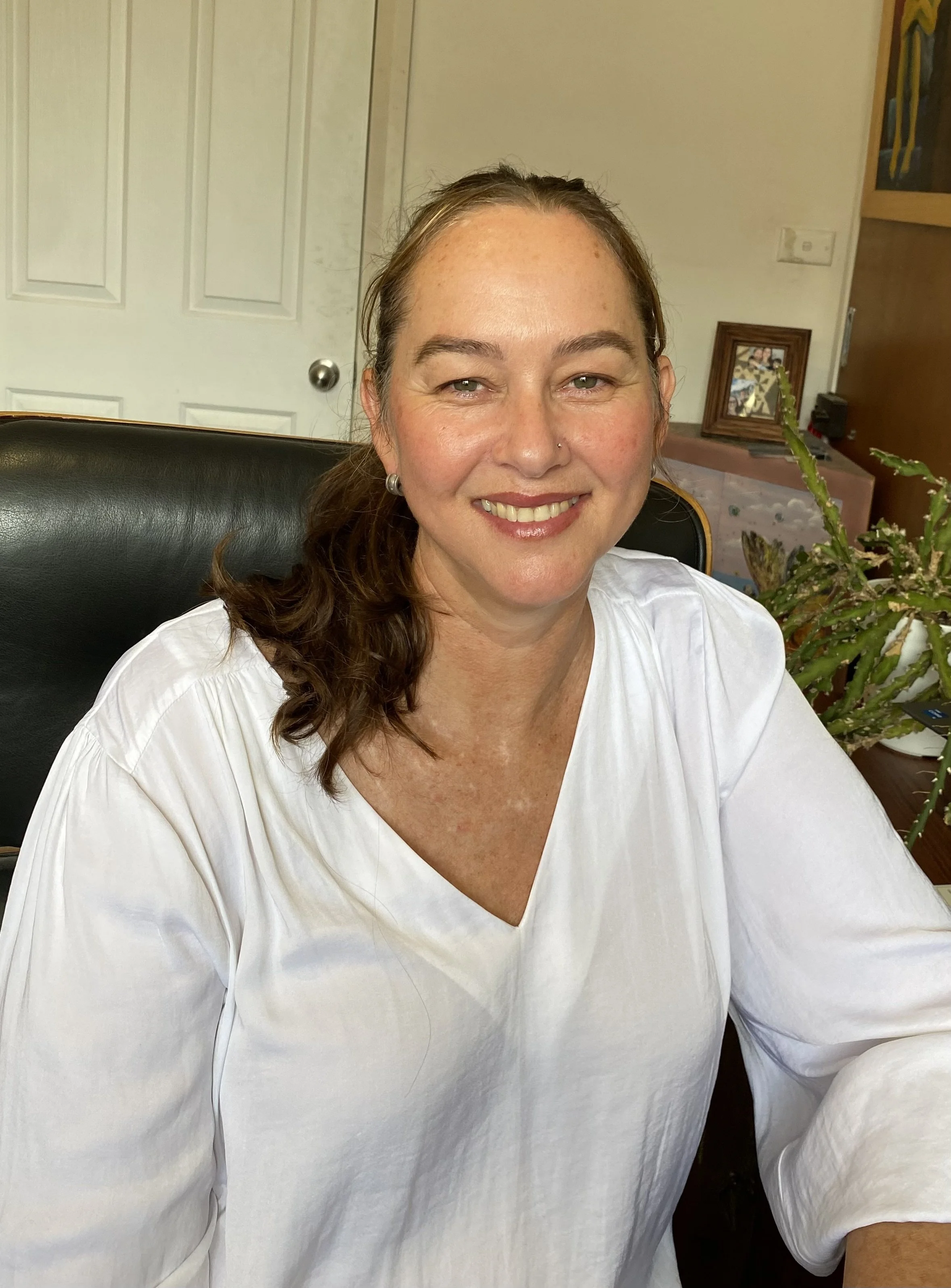A woman with brown hair wearing a white blouse, sitting at a desk in an office, smiling at the camera.