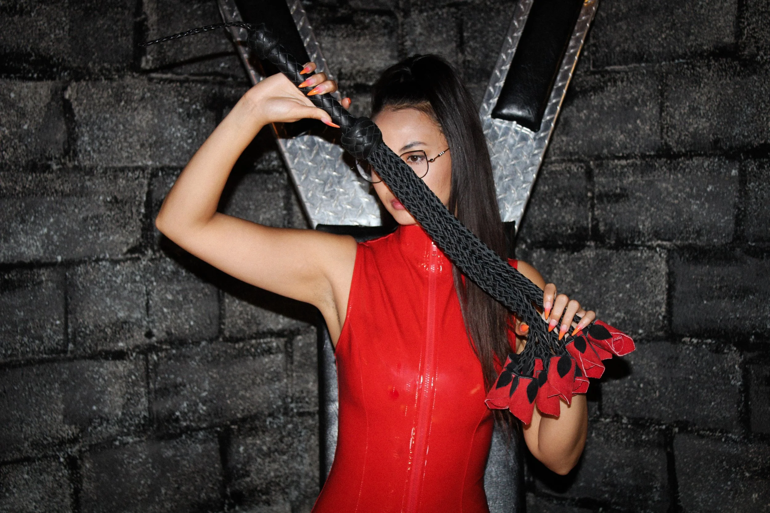 A woman in a red sleeveless outfit holding a braided whip with red and black tassels in front of a dark background with a metallic logo.