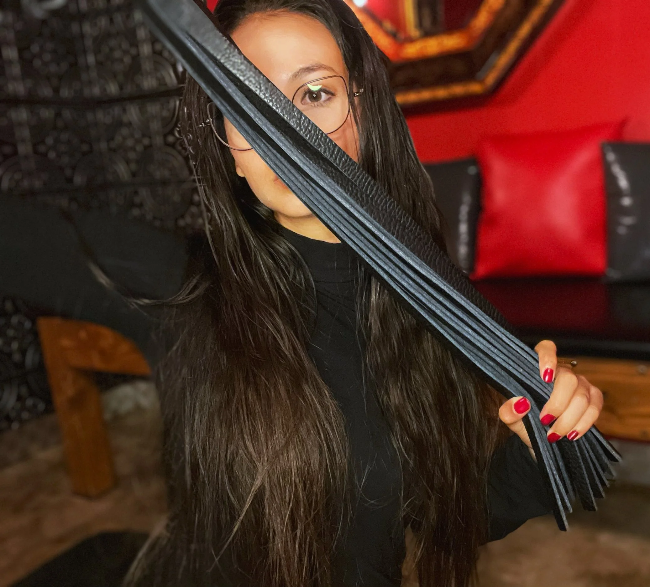 A woman with long brown hair, wearing glasses, holds a black fringe belt in front of her face, partially covering it, in a warmly decorated room with red and black cushions and wooden furniture.