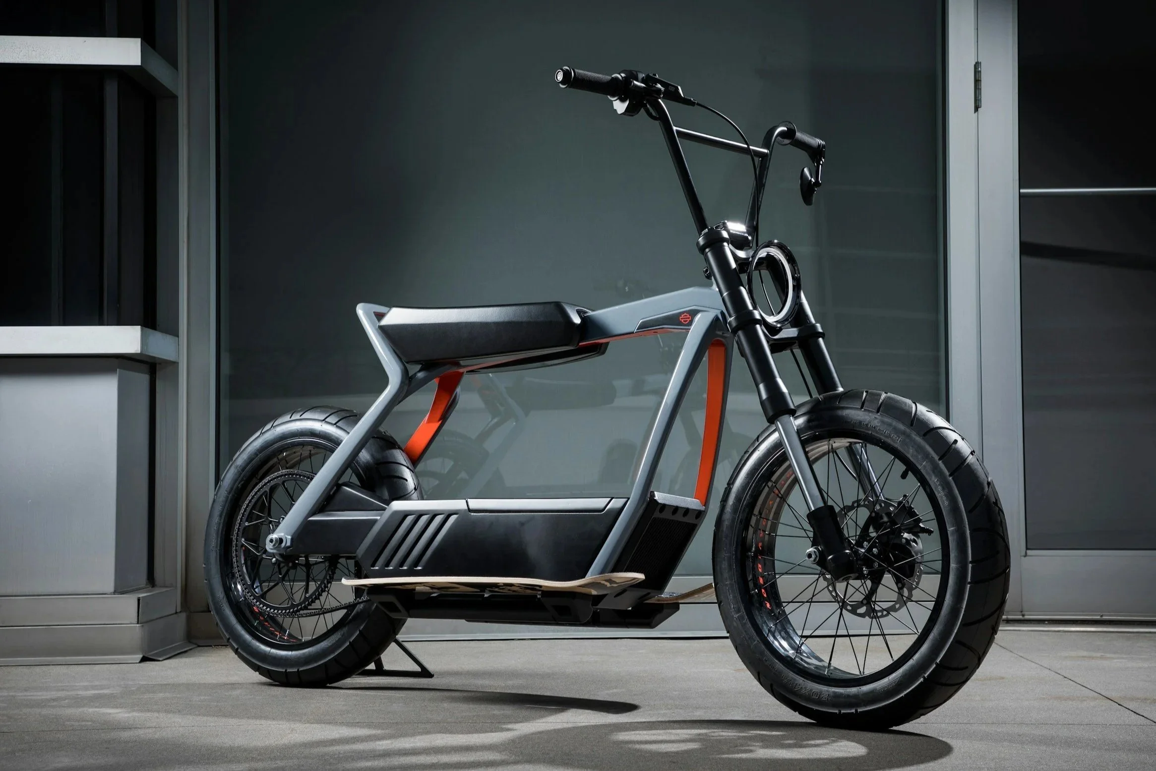 A sleek, modern electric motorcycle with a matte gray frame and black tires, parked in front of a dark wall and a glass door.