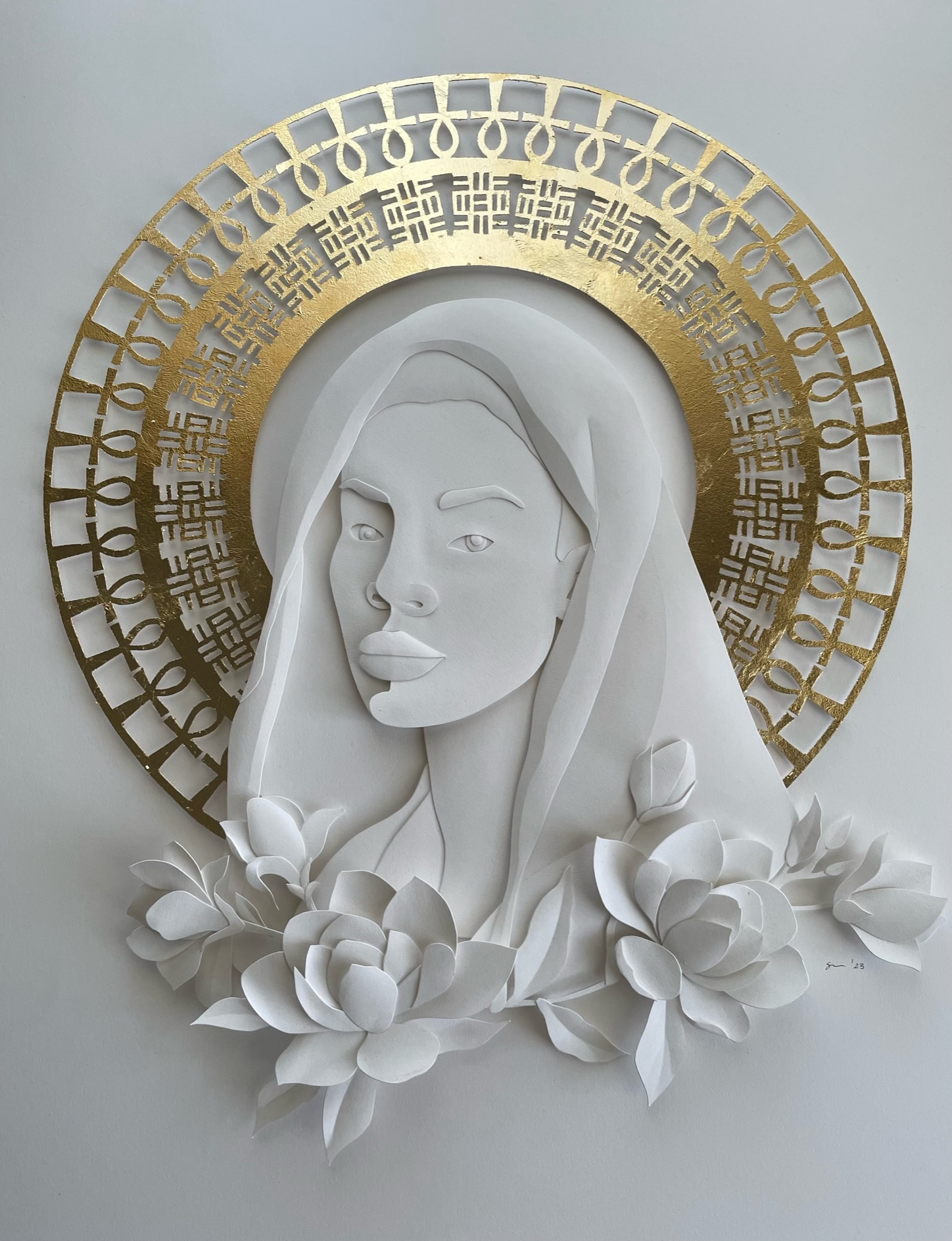 Paper art depicting a woman's face with long hair, surrounded by a gold halo and white floral elements.