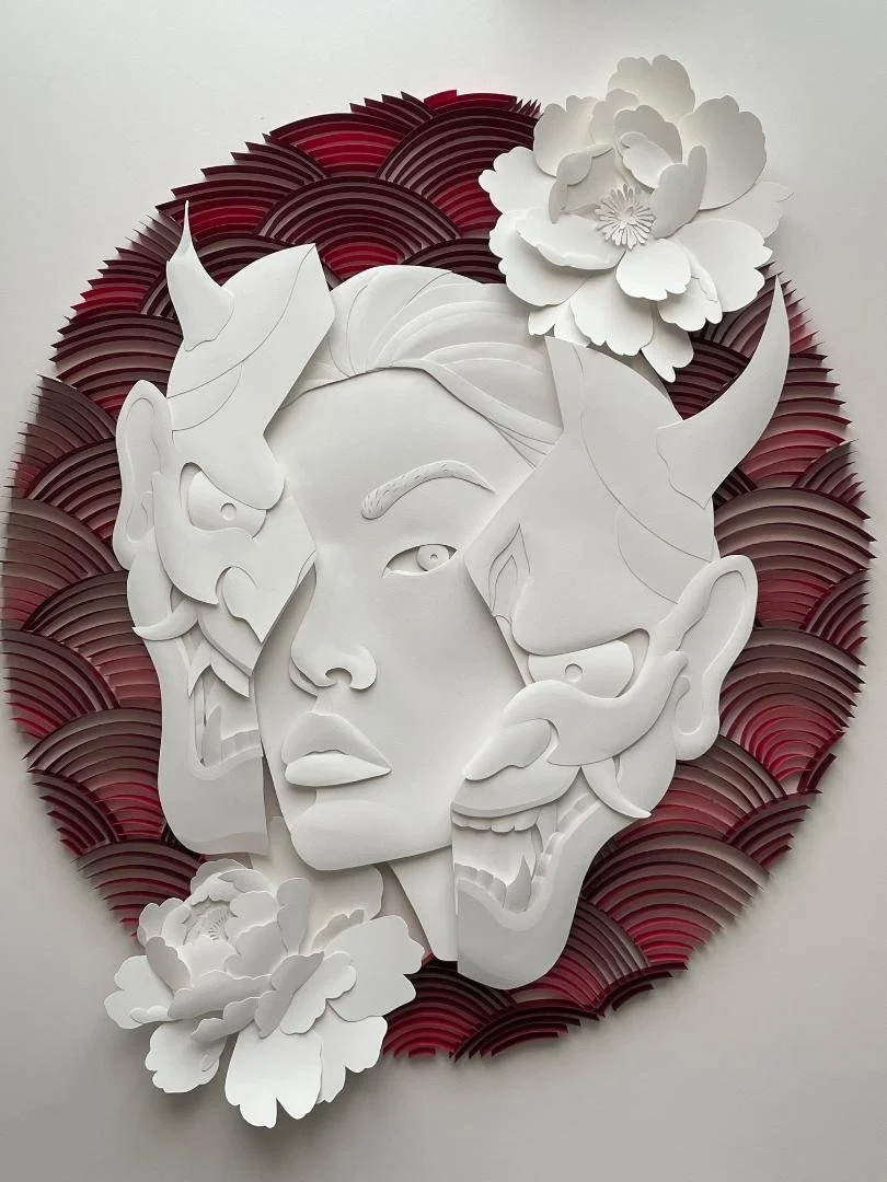 Papercraft artwork of a woman's face with oni mask and surrounded by flowers and a decorative red and black background. Traditional Japanese wave pattern forms circle paying tribute to the Japanese flag.