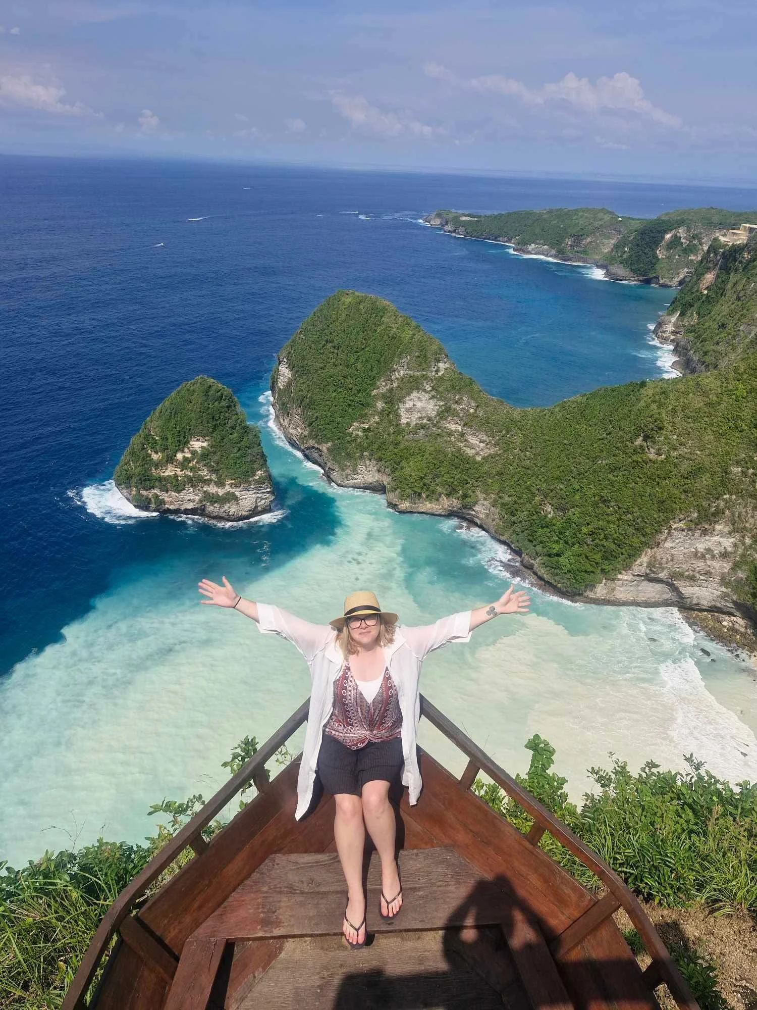 A woman sitting on a wooden platform with her arms outstretched, overlooking a tropical beach and green islands in the ocean.