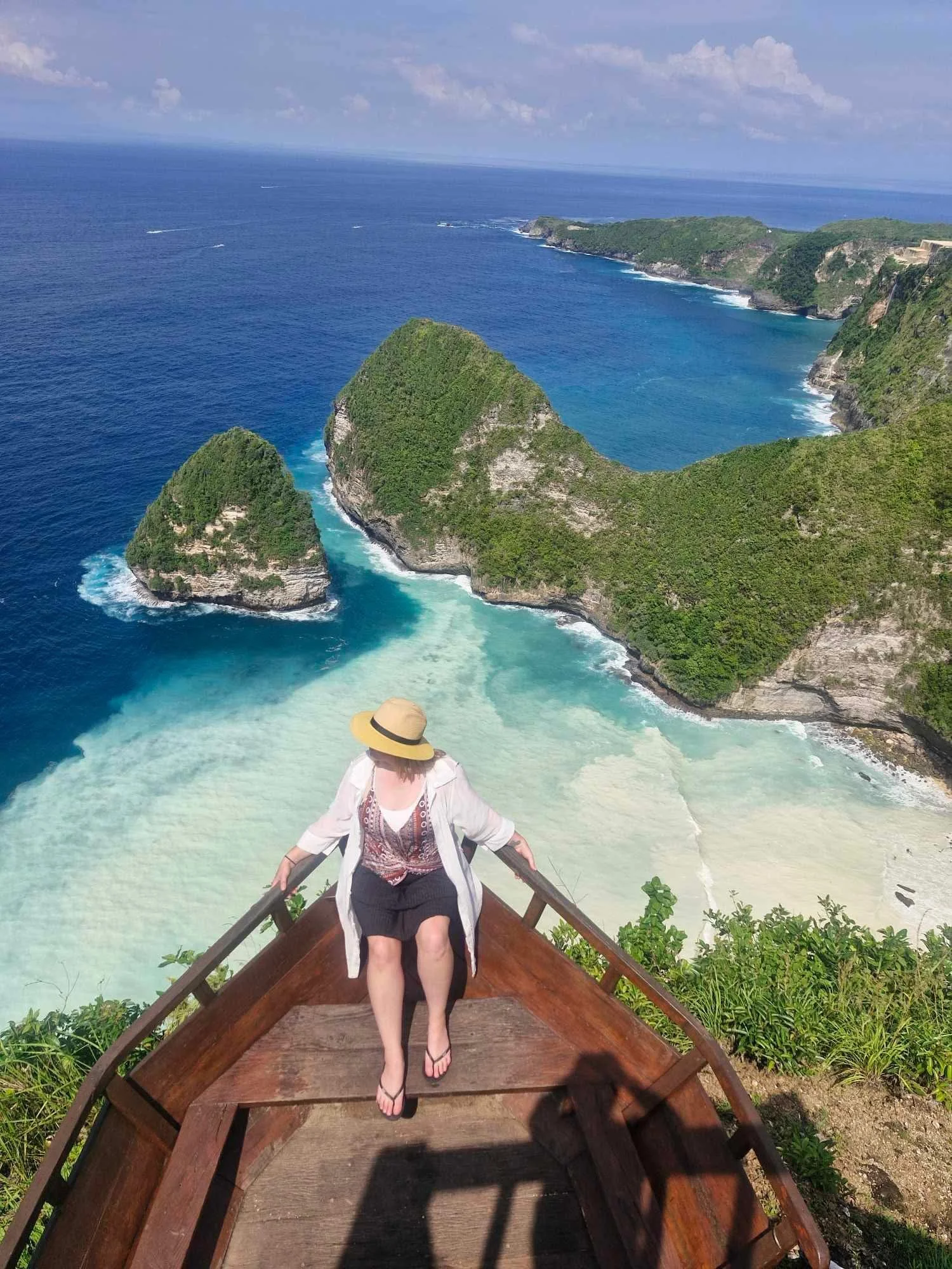 A woman sitting on a wooden boat dock, overlooking a lush, green coastline and blue ocean with cliffs, under a partly cloudy sky.