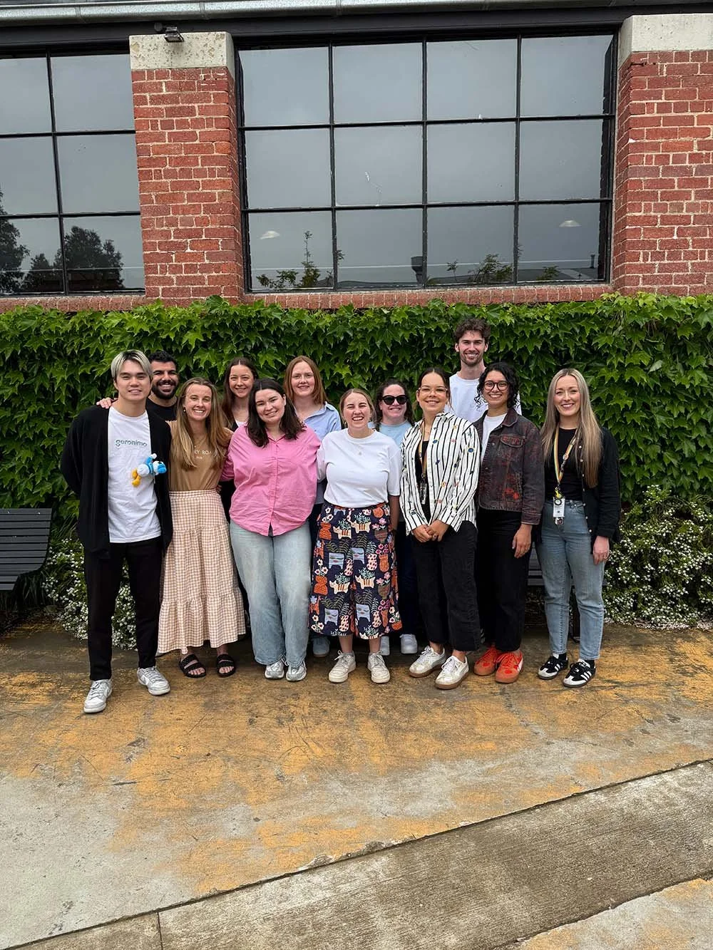 Group of ten diverse young adults standing outdoors in front of a green leafy hedge, with a brick and glass building behind them, smiling at the camera.