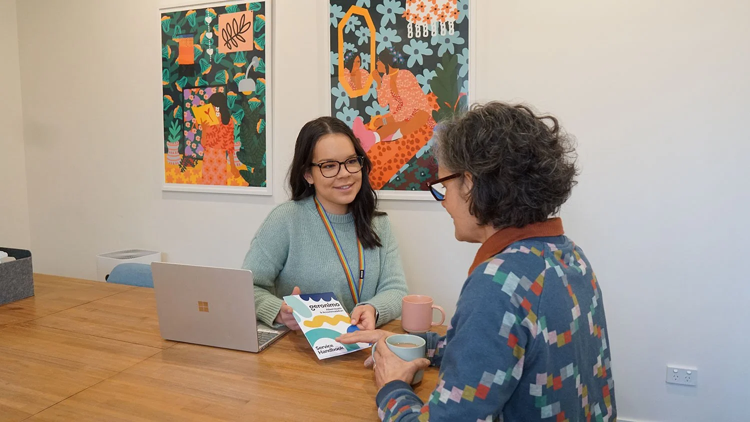Two women having a conversation at a wooden table with colorful artwork on the wall behind them. One woman has dark hair, glasses, and is wearing a light green sweater and a rainbow lanyard, holding a pamphlet. The other woman has gray curly hair, glasses, and is wearing a patterned blazer, holding two mugs.