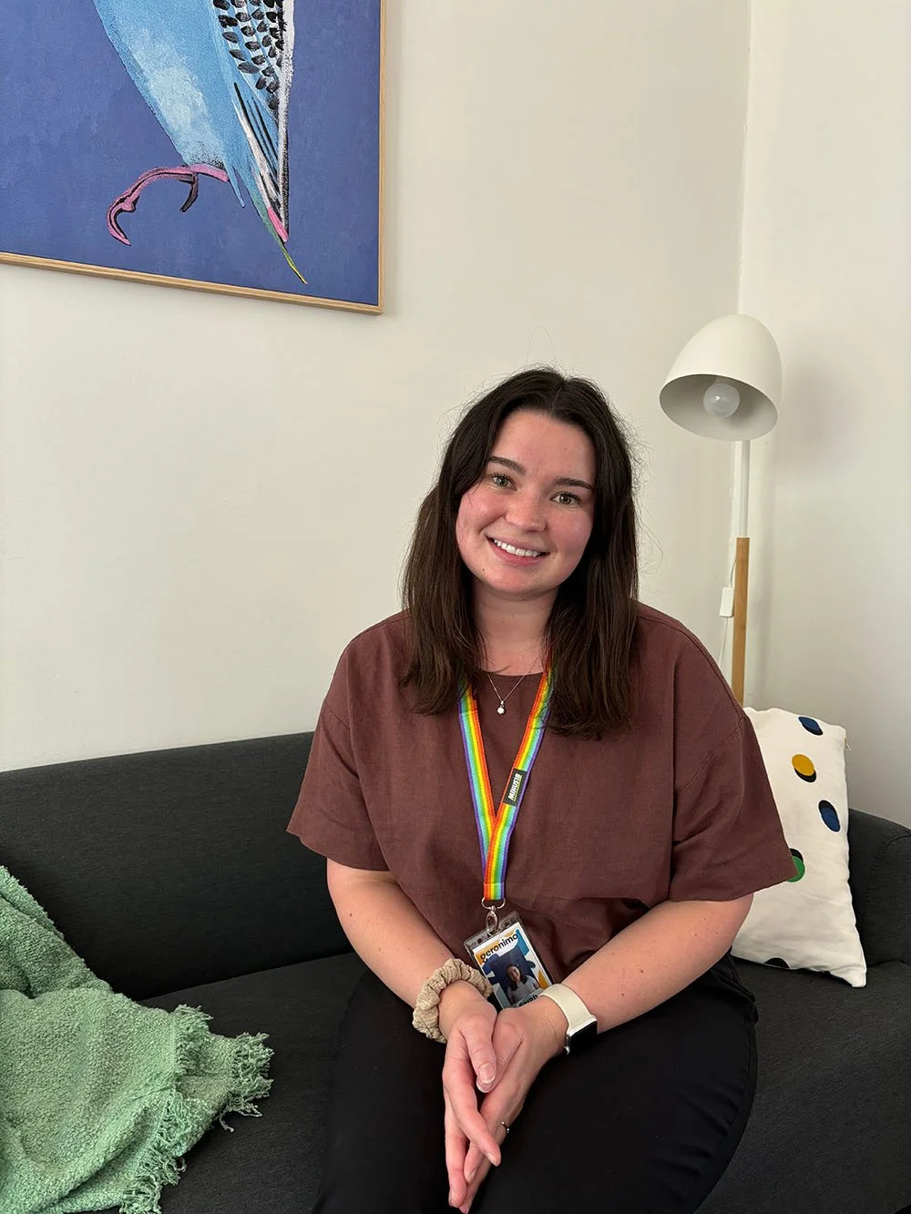 A young woman with dark brown hair, wearing a brown t-shirt, black pants, and a rainbow-colored lanyard, sitting on a black couch in a room decorated with colorful art and pillows, smiling at the camera.