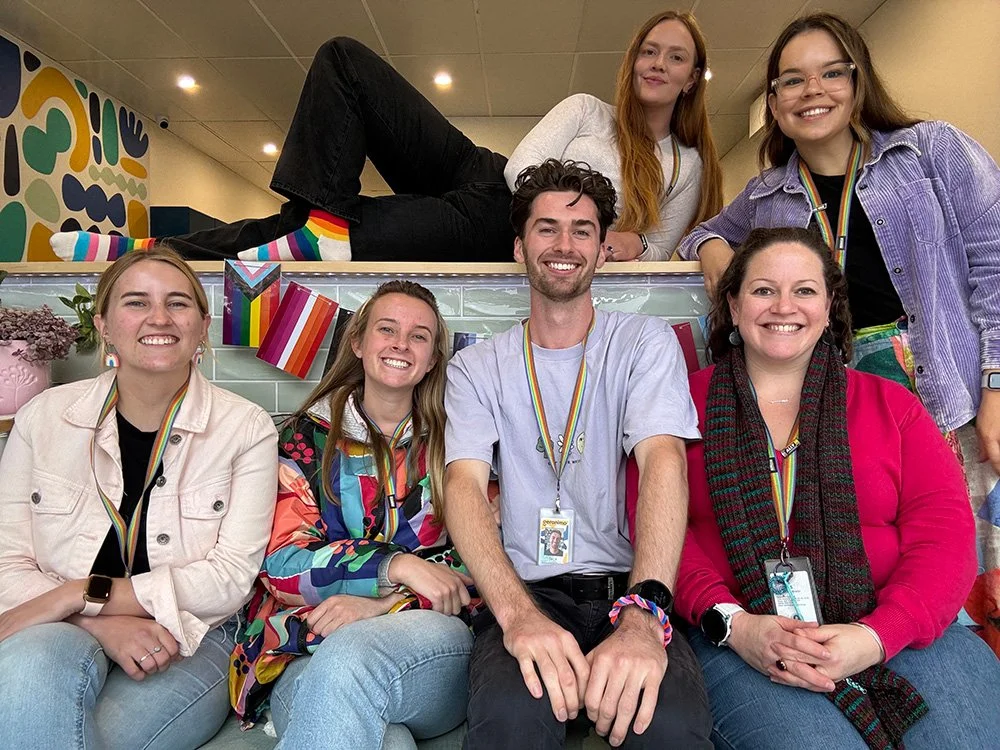 Group of seven young adults celebrating LGBTQ+ pride, wearing rainbow medals, smiling, with Pride flags and colorful decorations in the background.