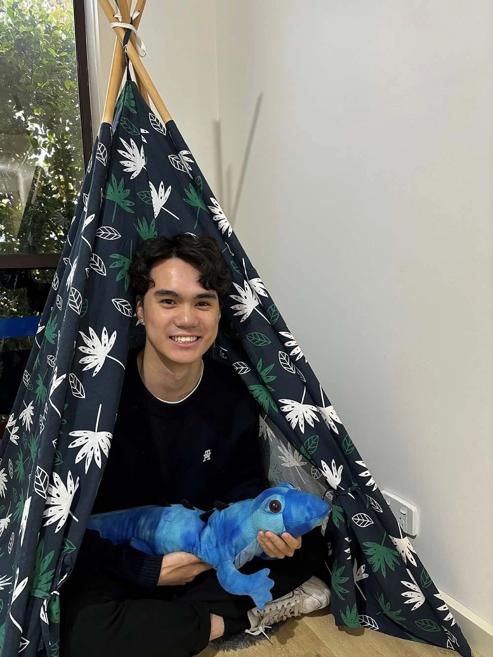 A young man sitting on the floor inside a fabric tent with a leafy pattern, holding a blue plush toy that looks like a lizard or dinosaur, smiling at the camera. There is a window with greenery outside to his left.