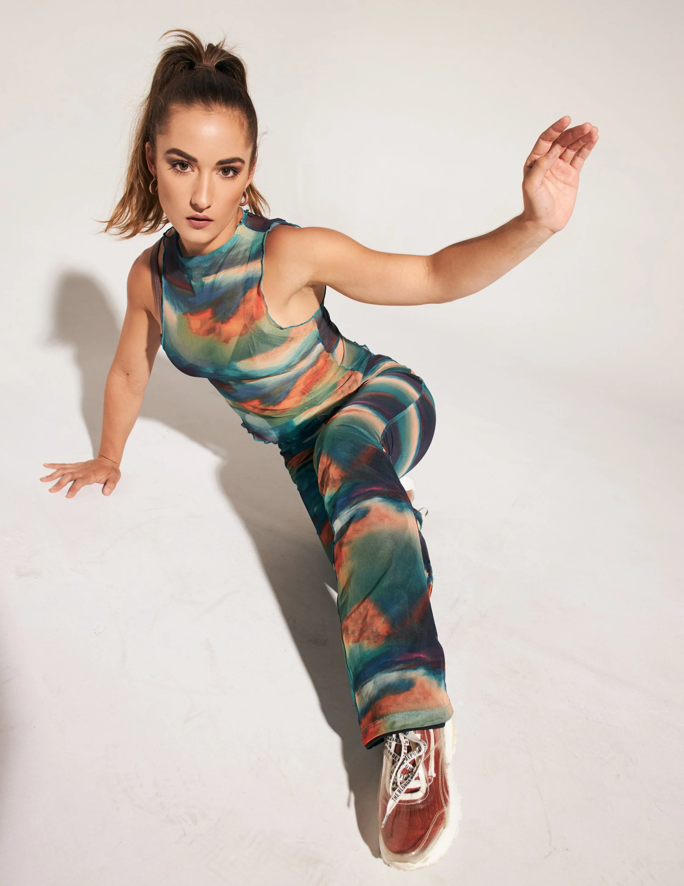 A woman in colorful, tie-dye athletic wear crouches on the floor with one arm extended and the other hand on the ground, against a plain white background.