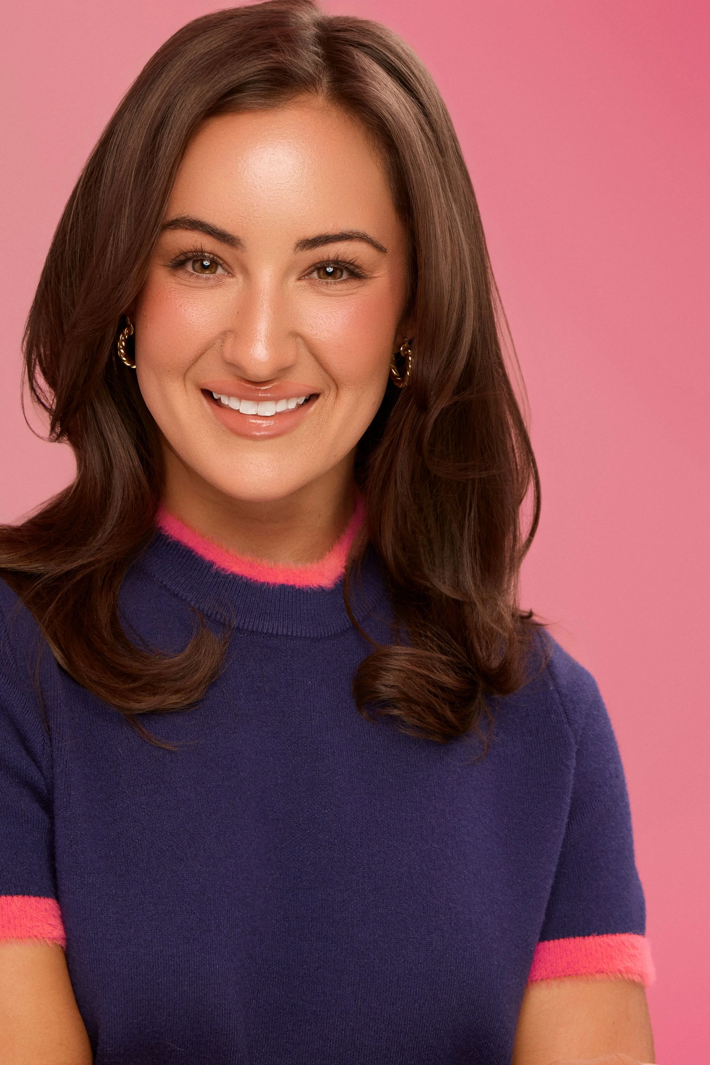 A woman smiling with light skin, long brown hair, wearing a dark blue sweater with pink trim, gold hoop earrings, against a pink background.