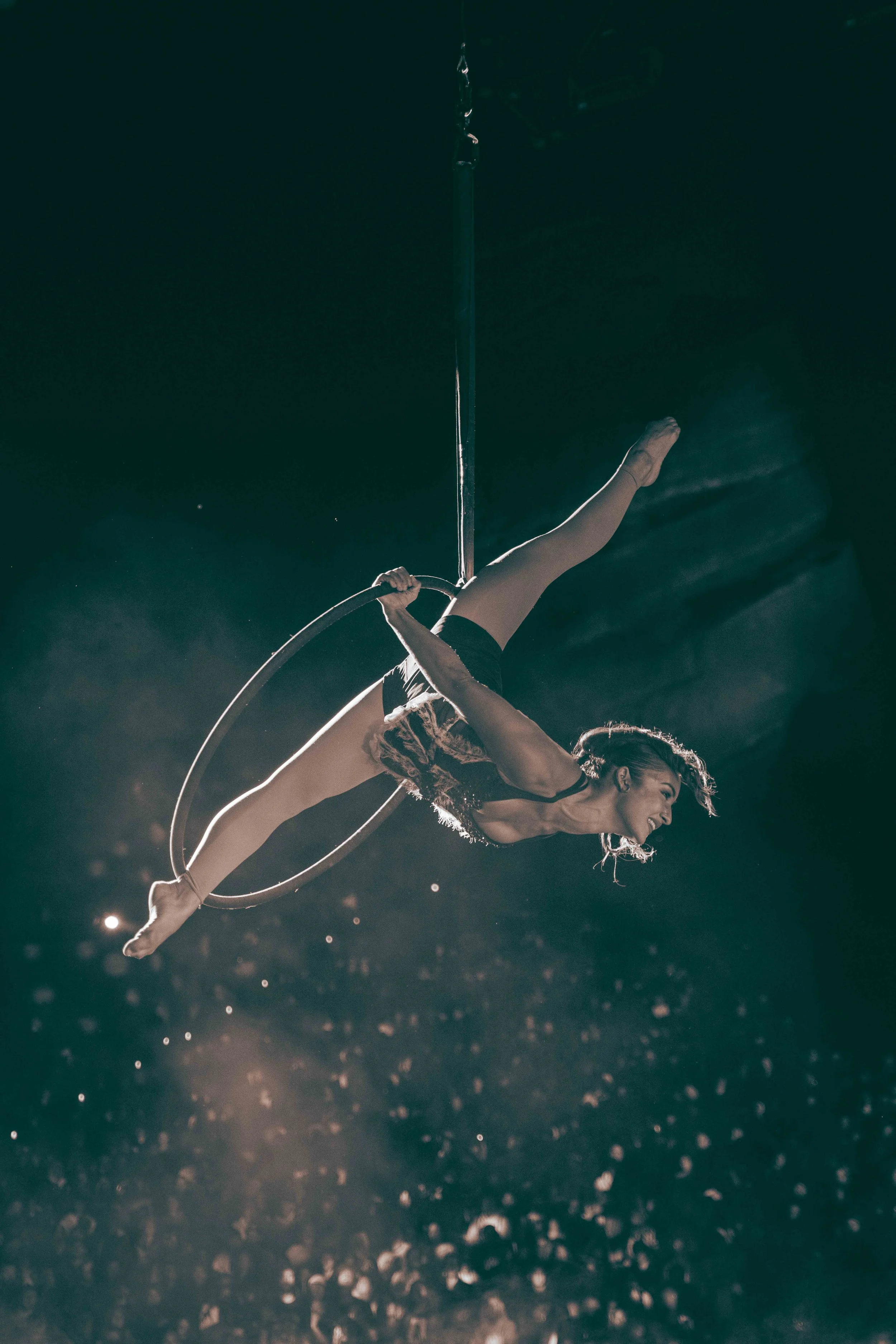A female aerialist performing a routine on an aerial hoop in a dimly lit theater, illuminated by stage lights.