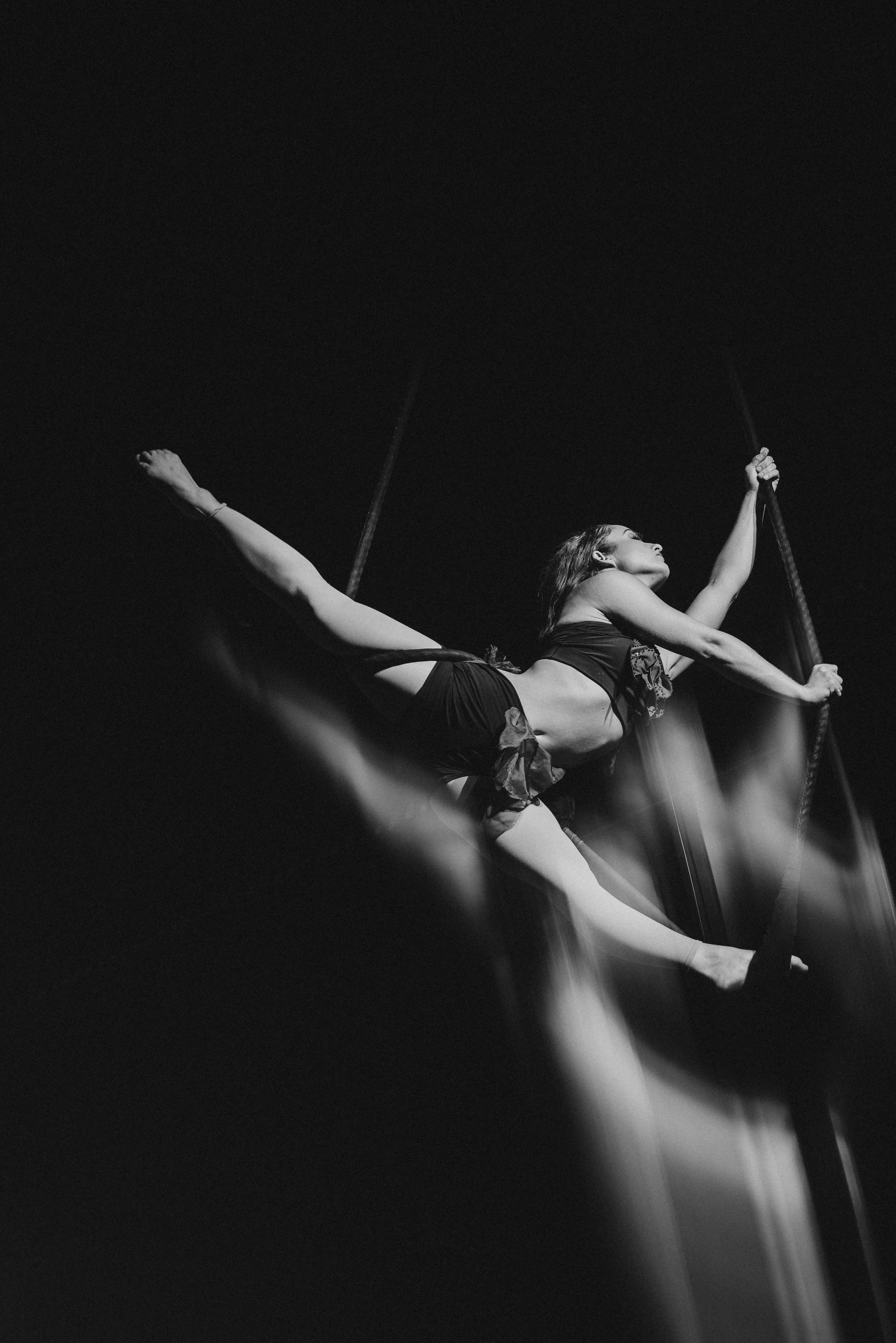 A female performer in a black costume poses dramatically on a swinging trapeze against a dark background.