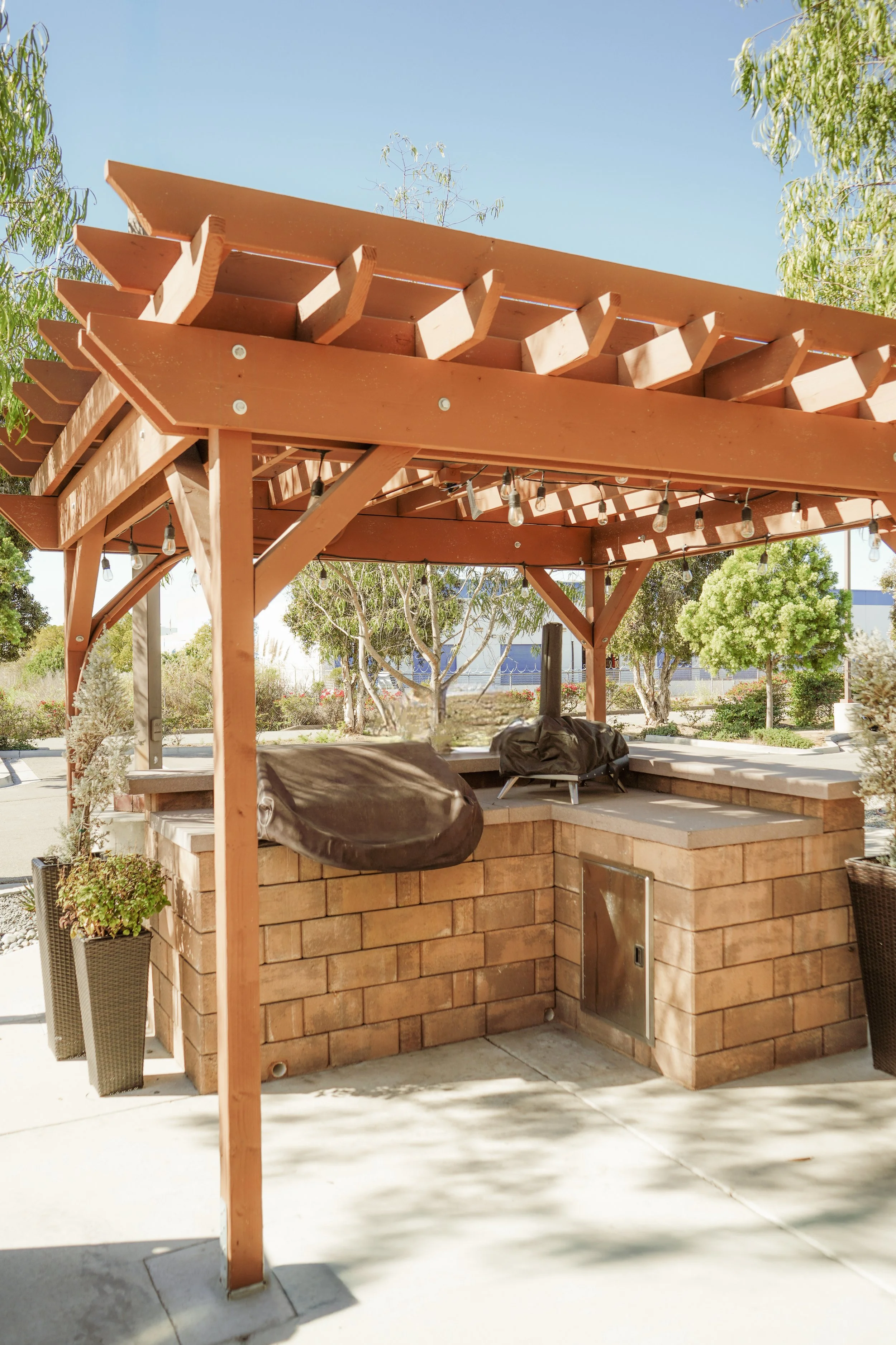 Outdoor brick barbecue grill under a wooden pergola with string lights, surrounded by trees and bushes.