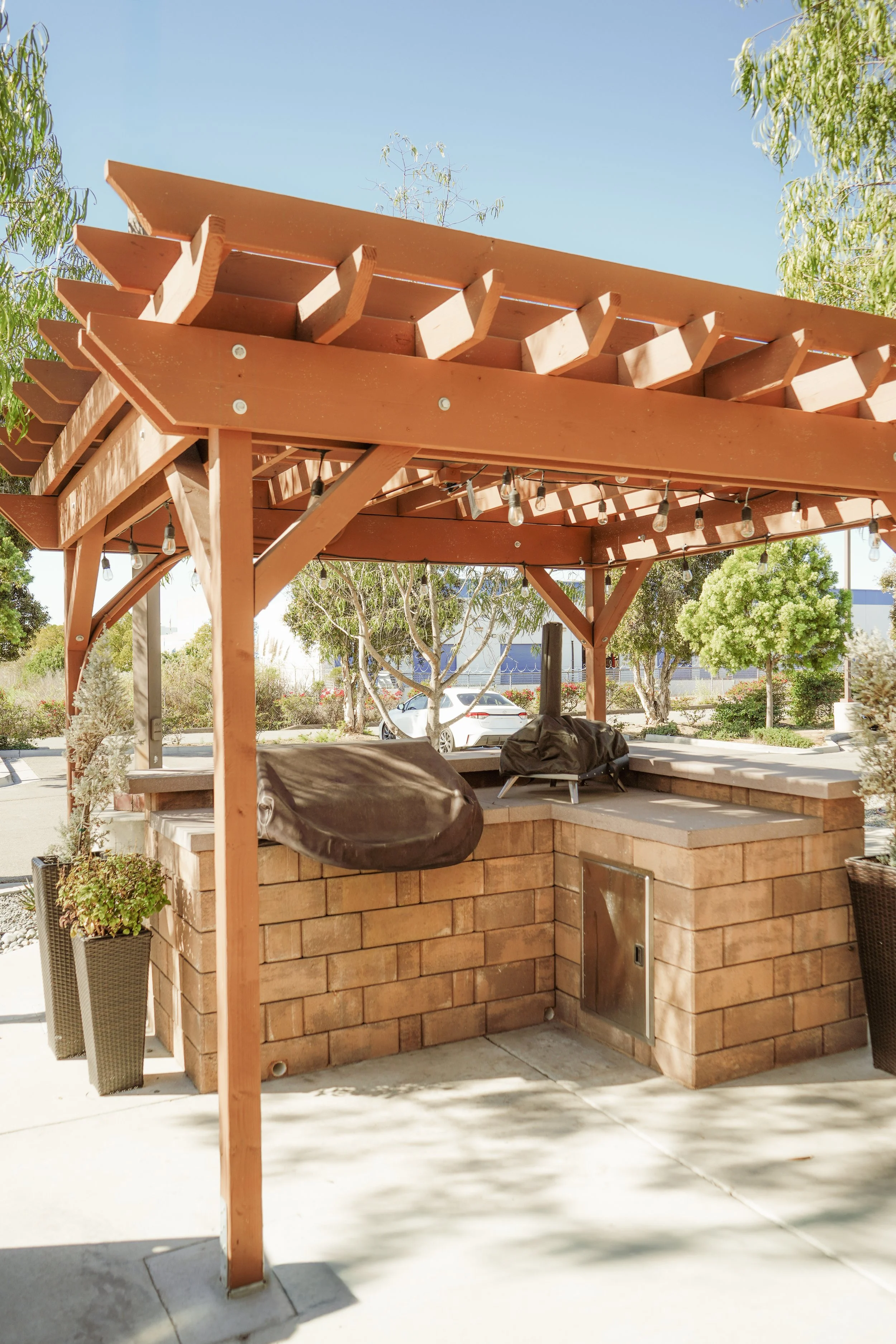 An outdoor brick bar with a wooden pergola and string lights, surrounded by trees and plants in a sunny setting.