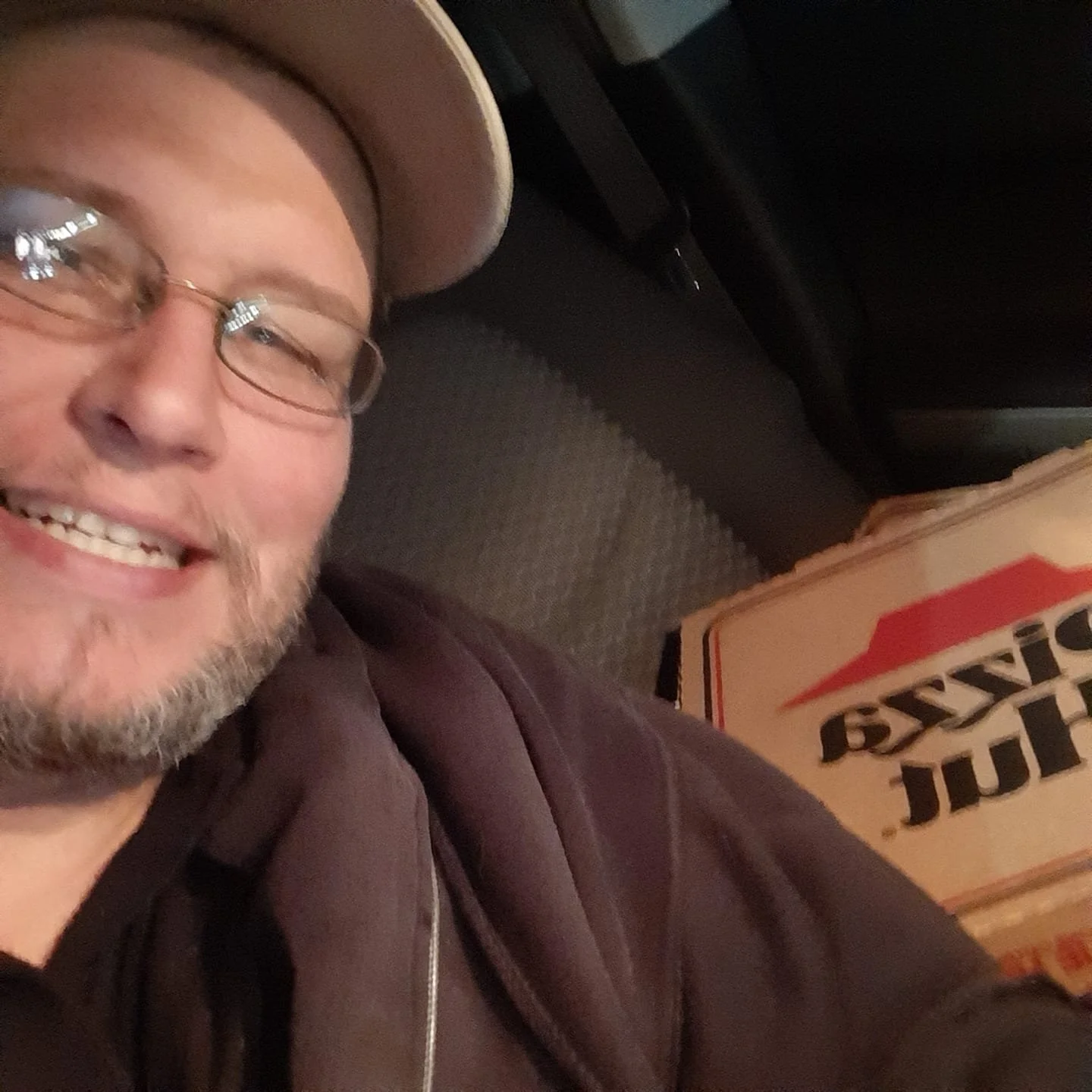 A smiling man wearing glasses and a beige cap, reclining on a couch with a pizza box beside him.
