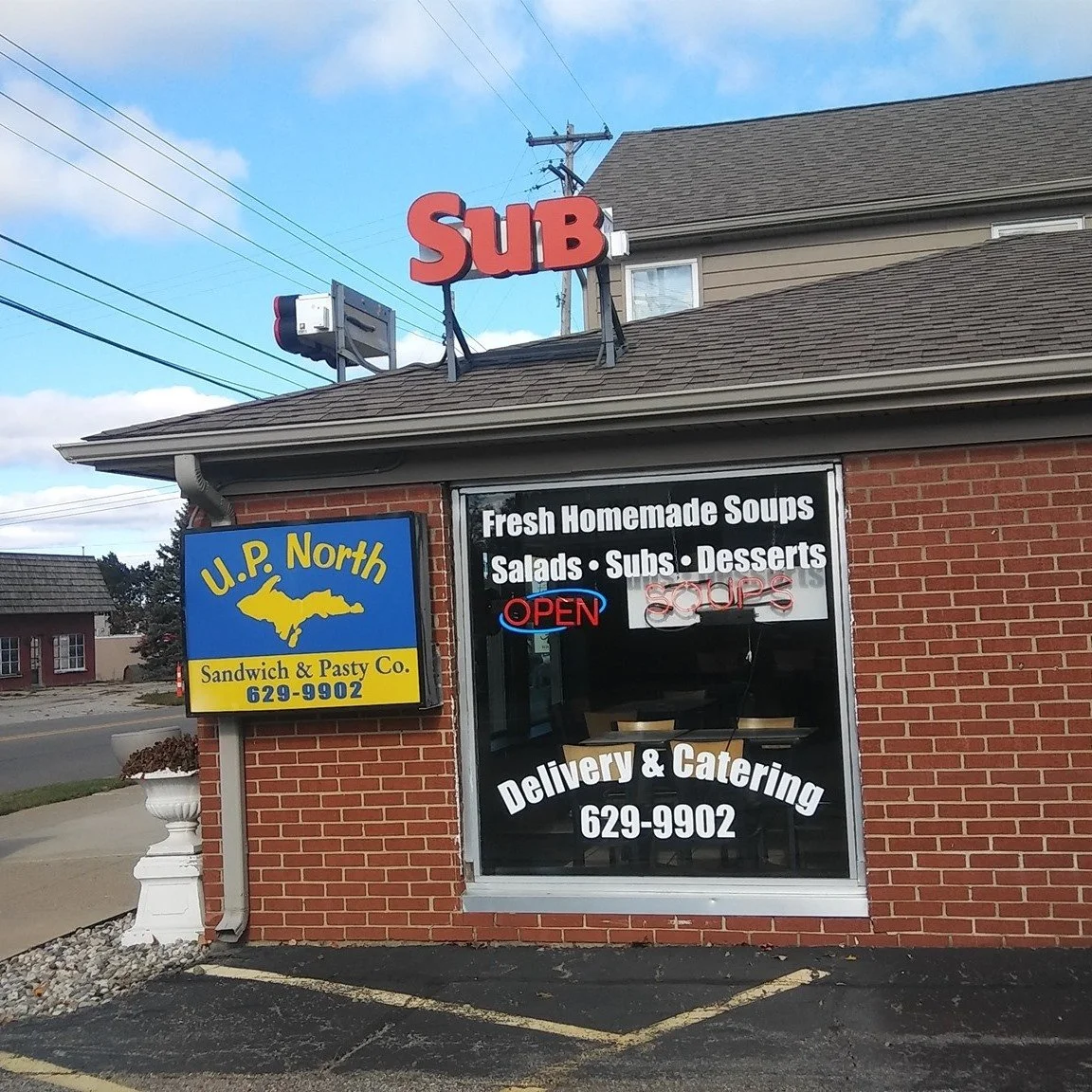 Front of a brick restaurant with signs advertising homemade soups, salads, subs, and desserts, including a neon 'OPEN' sign. An additional sign for U.P. North Sandwich & Pasty Co. is on the side of the building.