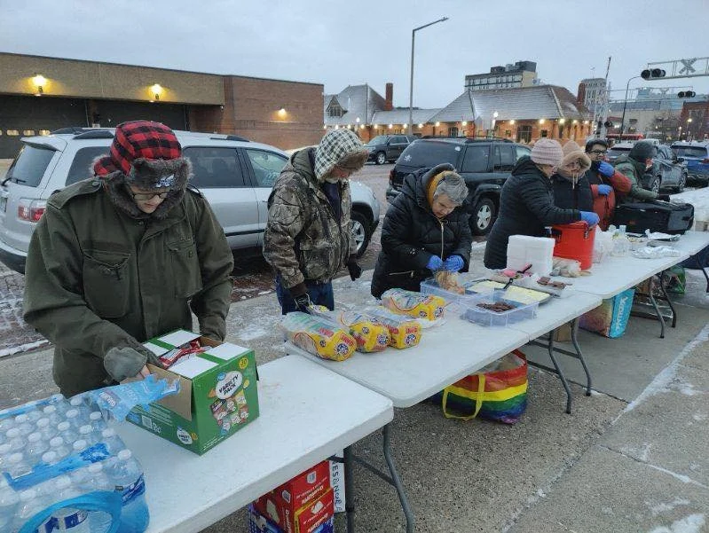 People standing behind a long table outdoors, serving and preparing food at a charity or community event in winter, with cars parked behind them.